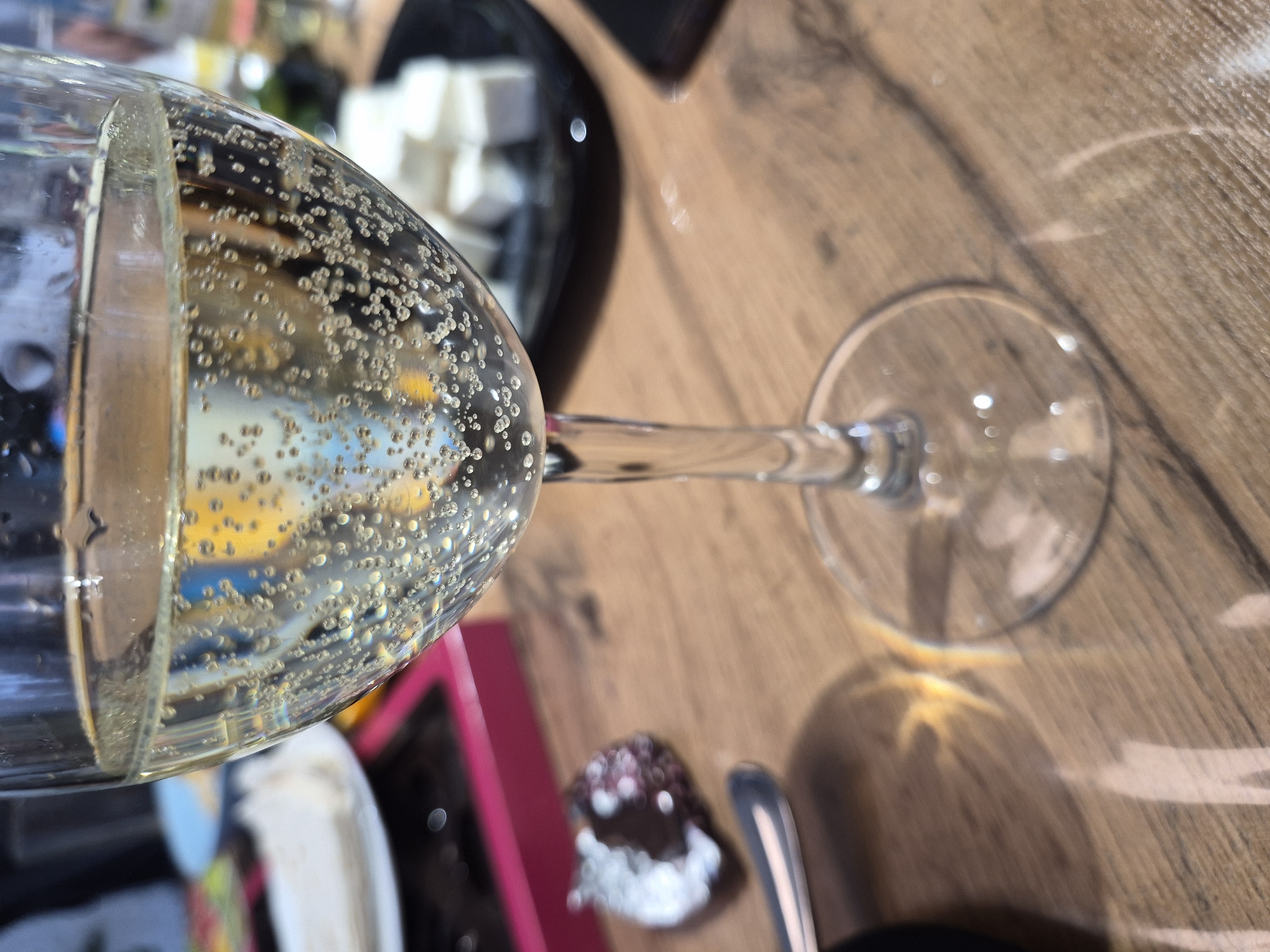 A close-up, slightly blurred, high-angle shot depicts a glass of sparkling wine or champagne filled with bubbles, situated on a wooden surface. To the left, a black plate contains white squares, possibly sugar cubes or candies. A chocolate-covered treat with white icing and a spoon are also visible near the bottom of the frame. The scene includes elements suggesting a celebratory or festive occasion. The location, Chaikovsky, Russia, is not directly evident in the image but has been requested contextually. No people are visible.
