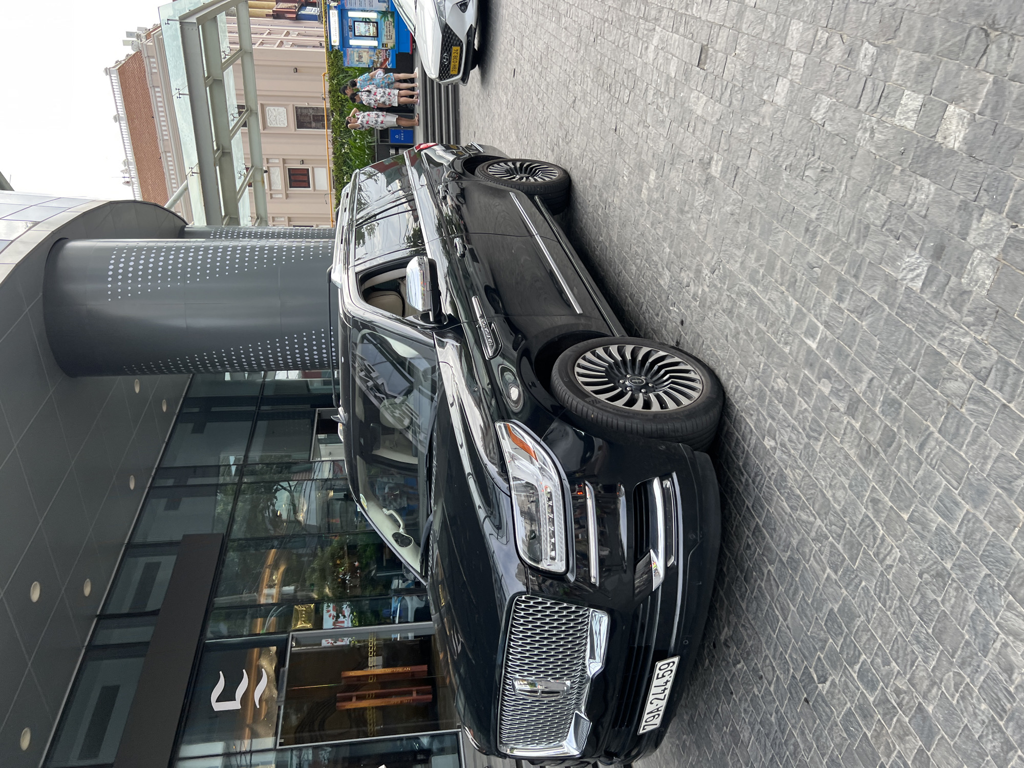 The image captures a black Lincoln Navigator parked on a stone-paved driveway in front of a modern building with large glass windows. The building appears to be a commercial establishment, possibly a hotel or a luxury store, as suggested by its architecture and the signage visible on the glass. In the background, a glimpse of a traditional building with a red-tiled roof can be seen, indicating a blend of modern and historical architecture in the city. Several people are visible in the distance, standing near a kiosk or information booth. The license plate on the Lincoln Navigator reads "79A-244.59", suggesting it is registered in Vietnam. The sky is overcast, indicating a possibly humid or rainy day. The scene is likely in Nha Trang, Vietnam, given the vehicle's license plate format and the architectural style observed.