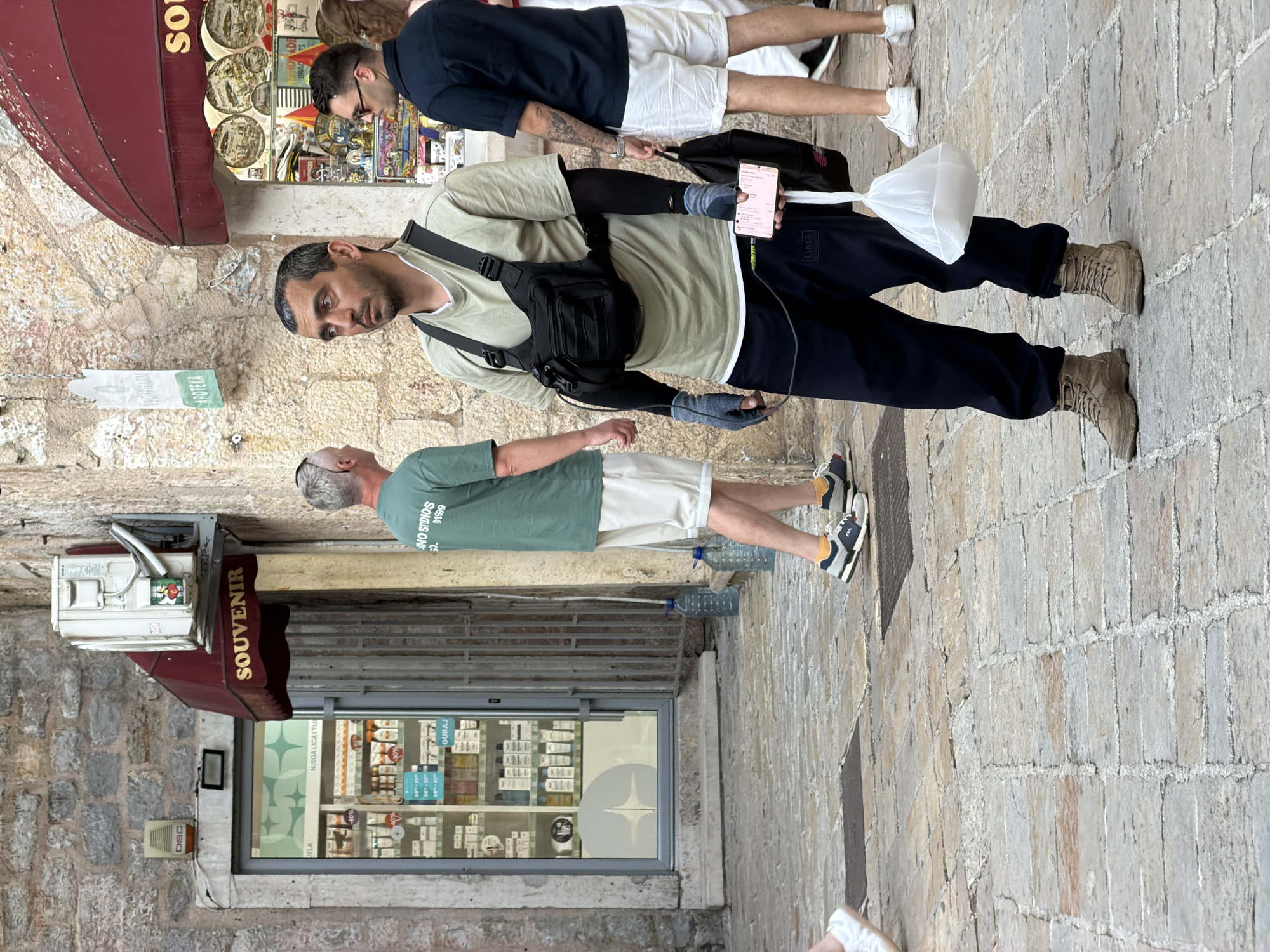 In the city of Budva, Montenegro, a man stands on a cobblestone street, facing right and looking to the side. He is wearing a light green t-shirt, dark blue sweatpants, and beige combat boots. A black tactical vest is worn over his t-shirt, with various pouches and straps. He has black compression sleeves on his arms and is wearing grey fingerless gloves. In his right hand, he holds a smartphone, and in his left hand, a white plastic bag containing an unidentifiable object.
Behind him, to the left, a man in a green t-shirt and white shorts walks away from the camera. Further left, a storefront labeled "DSC" with a security alarm is visible above a doorway. Adjacent to this, a shop with a red awning displays "SOUVENIR" in gold lettering and showcases various souvenir items in its window. To the right of the souvenir shop, another man in a dark blue short-sleeved shirt and white shorts is seen walking, also looking to the side. The buildings in the background are constructed from rough stone.