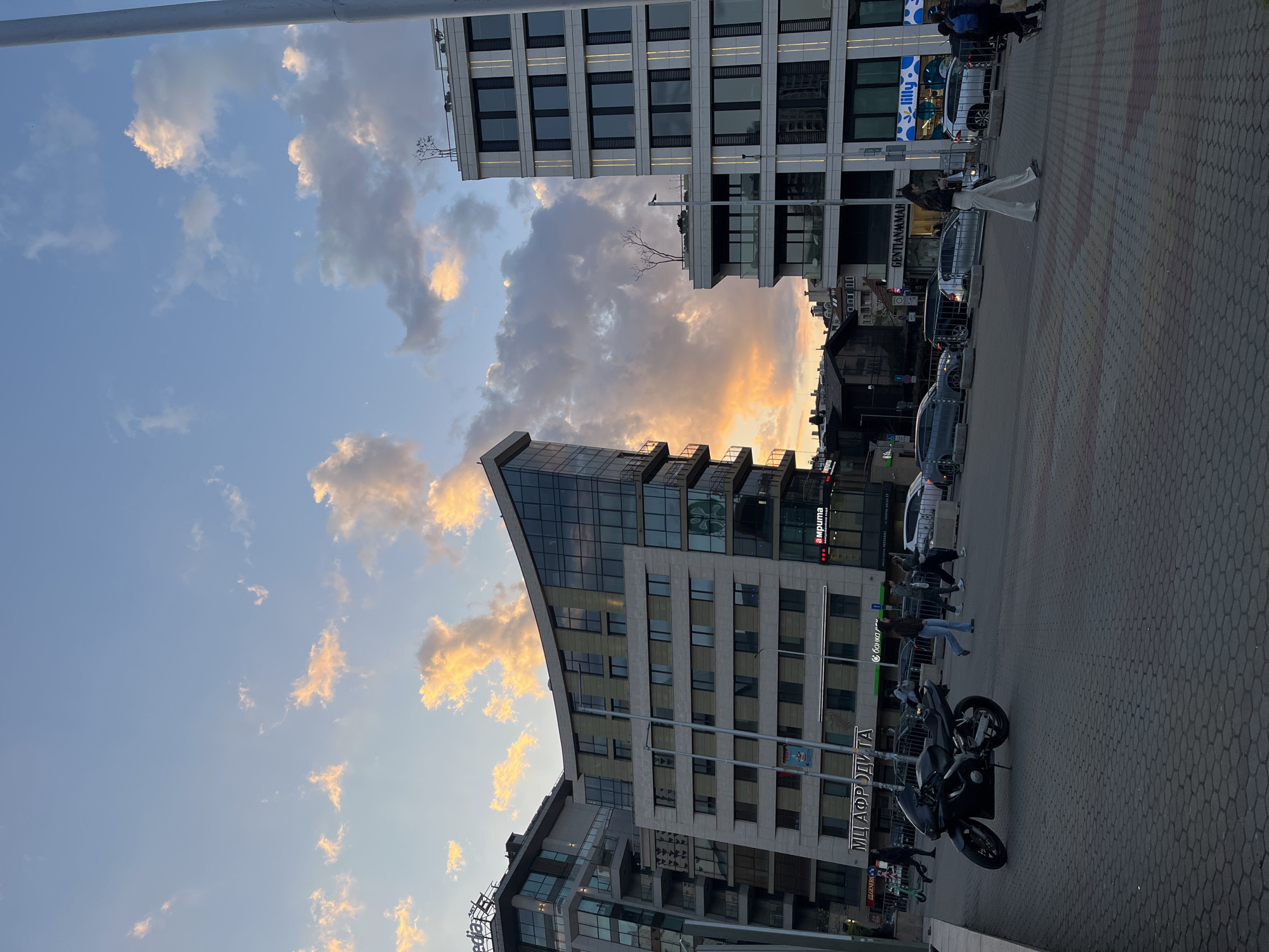 The image captures an urban scene in Sofia, Bulgaria. The sky is a mix of blue and clouds, with some clouds illuminated by the setting sun, casting an orange hue. Several modern buildings flank the scene, featuring glass and concrete architecture. Along the street level, people are walking and interacting in front of the buildings. A motorcycle is parked on the paved sidewalk in the foreground. Street signage is visible, with "МЛАФРОДІТА" prominently displayed on a building. Cars are parked along the street, further indicating the urban setting. Pedestrians are moving along the sidewalk. The overall atmosphere suggests a late afternoon or early evening.