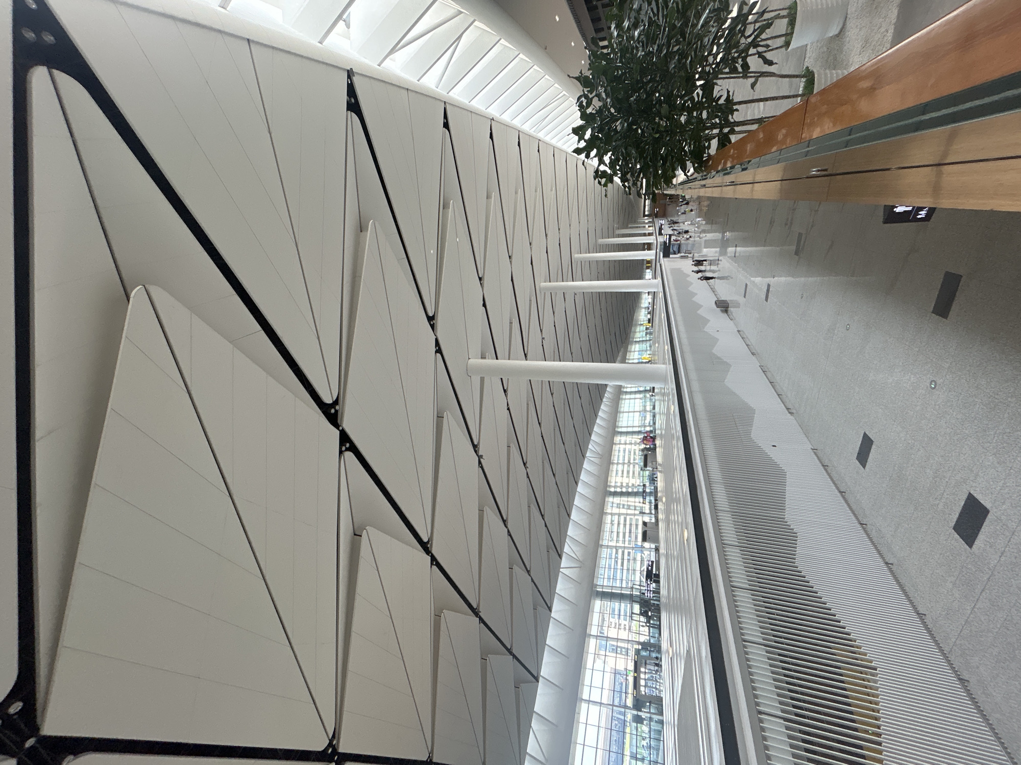 This is an interior, eye-level shot of a modern airport terminal in Jianyang City, China. The ceiling is a striking architectural feature, composed of a series of white, angled panels separated by dark frames, creating a visually dynamic pattern that recedes into the distance. Natural light streams in through large windows along the left side of the terminal, illuminating the spacious, polished floor. The floor reflects the overhead lights and the architectural elements.
Along the right side of the image, a lush indoor plant in a white pot adds a touch of greenery. A wooden railing with a glass barrier separates the viewpoint from the main concourse below. On the concourse level, several people are walking, appearing as small figures in the vast space, suggesting a bustling but orderly environment. The concourse features a white, perforated barrier with a wavy pattern. White pillars support the high ceiling, spaced evenly throughout the terminal. The overall impression is one of spaciousness, modern design, and efficient transit.