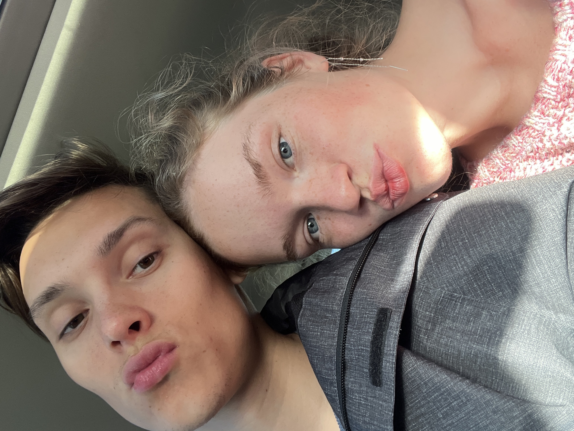 Two people are in close proximity; a woman leans her head on the shoulder of a man. The woman has fair skin with freckles and light blue eyes. She has light brown hair. She is wearing a pink and white knit shirt. The man has dark brown hair. They are both pouting at the camera. They are indoors with a gray surface visible in the background. Glavati, Montenegro, is not evident in the image.