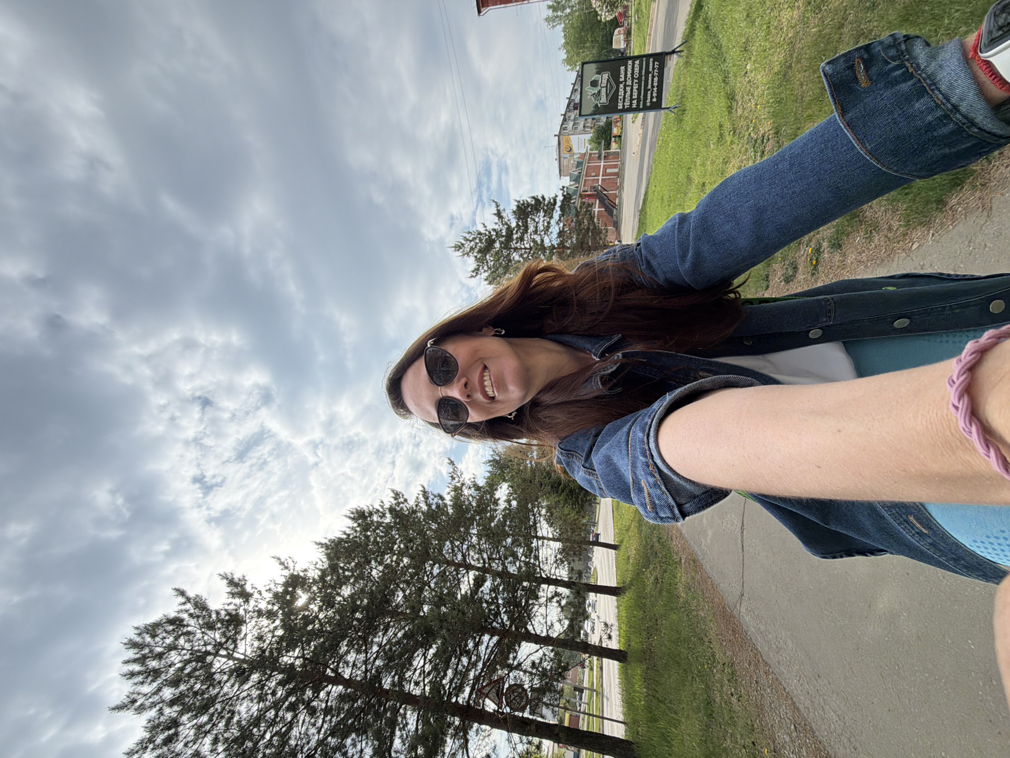 The image captures a selfie of a woman with long brown hair wearing sunglasses and a denim jacket. She is smiling. The background shows a cloudy sky and a street scene. Along the street are trees, grass, and buildings. There is a sign in the background. Based on the content of the sign in the background, the location is Birobidzhan, Russia.
