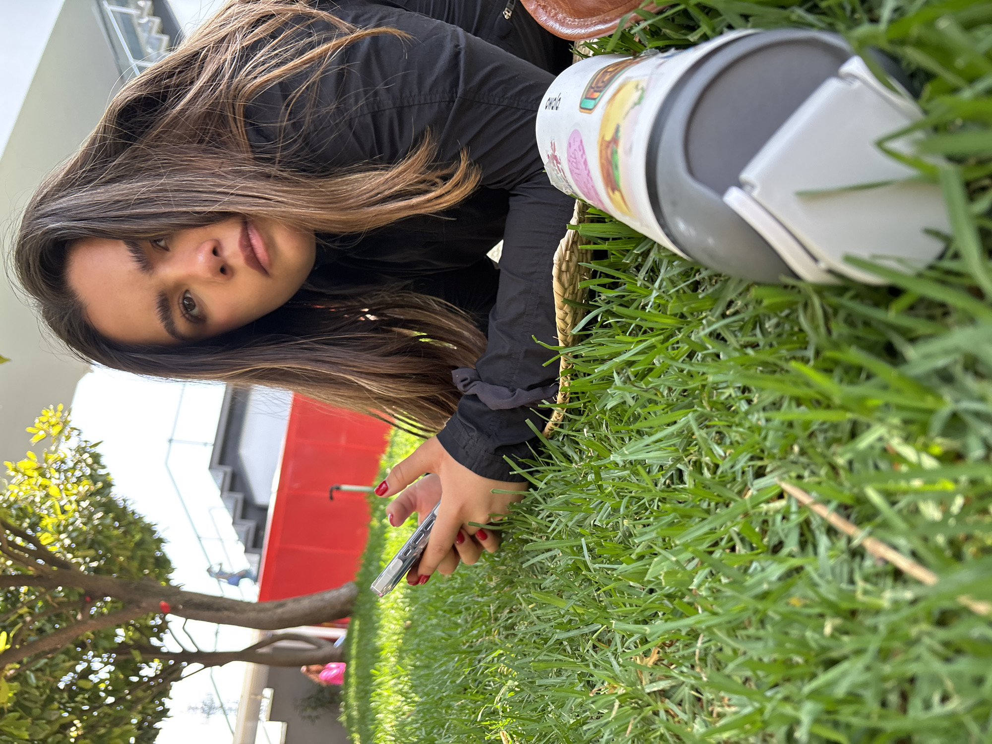 A young woman lies on a lawn with an open beverage container in front of her, holding a cell phone in her left hand. The location is outdoors in Mexico City, Mexico. The subject is lying at an angle to the camera. She has long, light brown hair and is wearing a black jacket. Her nails are painted red. She is holding a silver cell phone. The woman is close to a metal tumbler laying in the grass. The beverage container is lying on its side, with a silver lid open. A woven basket lies nearby. Behind the woman is a tree, and beyond that is a building with a red wall and stairs. The ground is covered in green grass. The light suggests it is daytime.