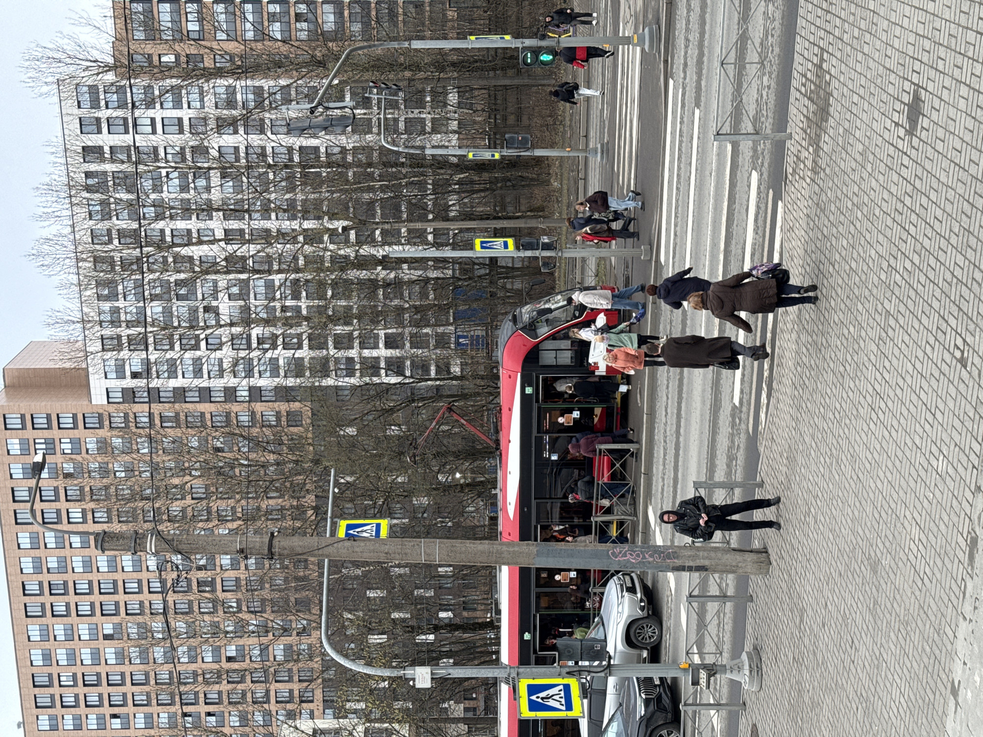A tram passes through a crosswalk in Saint Petersburg, Russia. Pedestrians are crossing the street. There are tall buildings in the background and trees. A crosswalk sign is visible with a yellow triangle and a blue image of a pedestrian. The crosswalk has white lines on the street, and the sidewalk is made of gray brick. Several cars are parked along the side of the road next to the tram. A streetlamp illuminates the scene. The traffic lights are green.