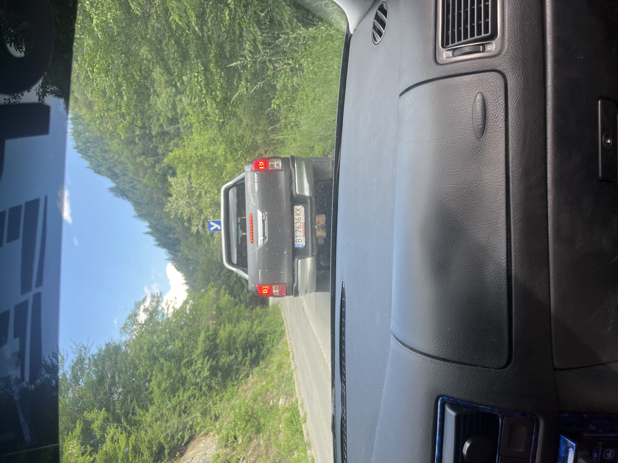 The image is taken from inside a car looking forward through the windshield. A dark-colored pickup truck is visible ahead, driving on a paved road that curves to the left. The truck has a license plate that reads "BT 7636 KK". A blue road sign with a white symbol is visible above the truck. The road is bordered by lush green trees and vegetation on both sides, with a steep, rocky incline on the left. The sky is blue with scattered clouds. The foreground of the image shows the dashboard of the car, including an "AIRBAG" label and an air vent. The scene appears to be in a rural, mountainous area.