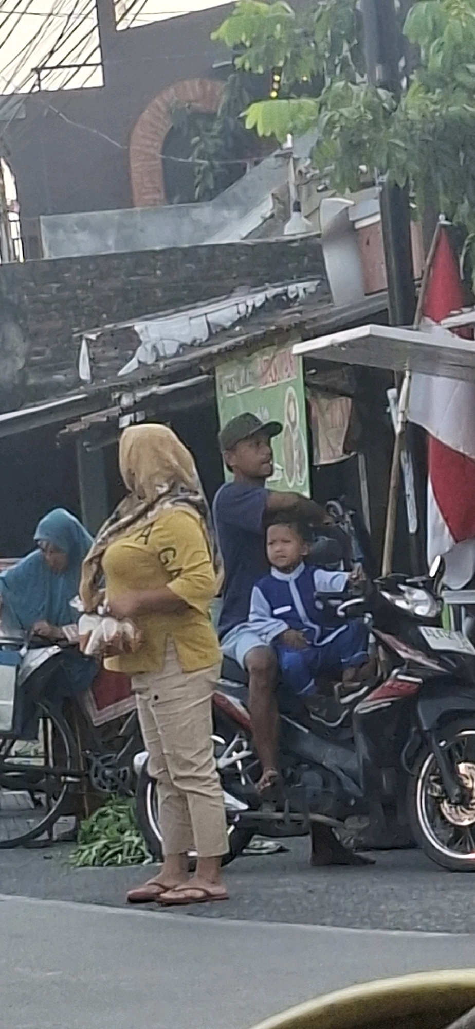 In Desa Dibal, Indonesia, a street scene unfolds with several individuals and vehicles. A woman wearing a yellow hijab and a yellow t-shirt with an orange motif stands on the left, holding what appears to be food packaging. She is wearing beige pants and red flip-flops. To her right, a man in a dark blue t-shirt and a green baseball cap is seated on a motorcycle, holding a young boy on his lap. The boy is dressed in a blue and white outfit. Another person, partially visible, is seated on a bicycle to the far left, wearing a blue hijab. The motorcycle is black with red accents and a license plate. In the background, a makeshift stall or shop is visible with a green banner and various items displayed. The environment suggests an outdoor urban or semi-urban setting with buildings and foliage.