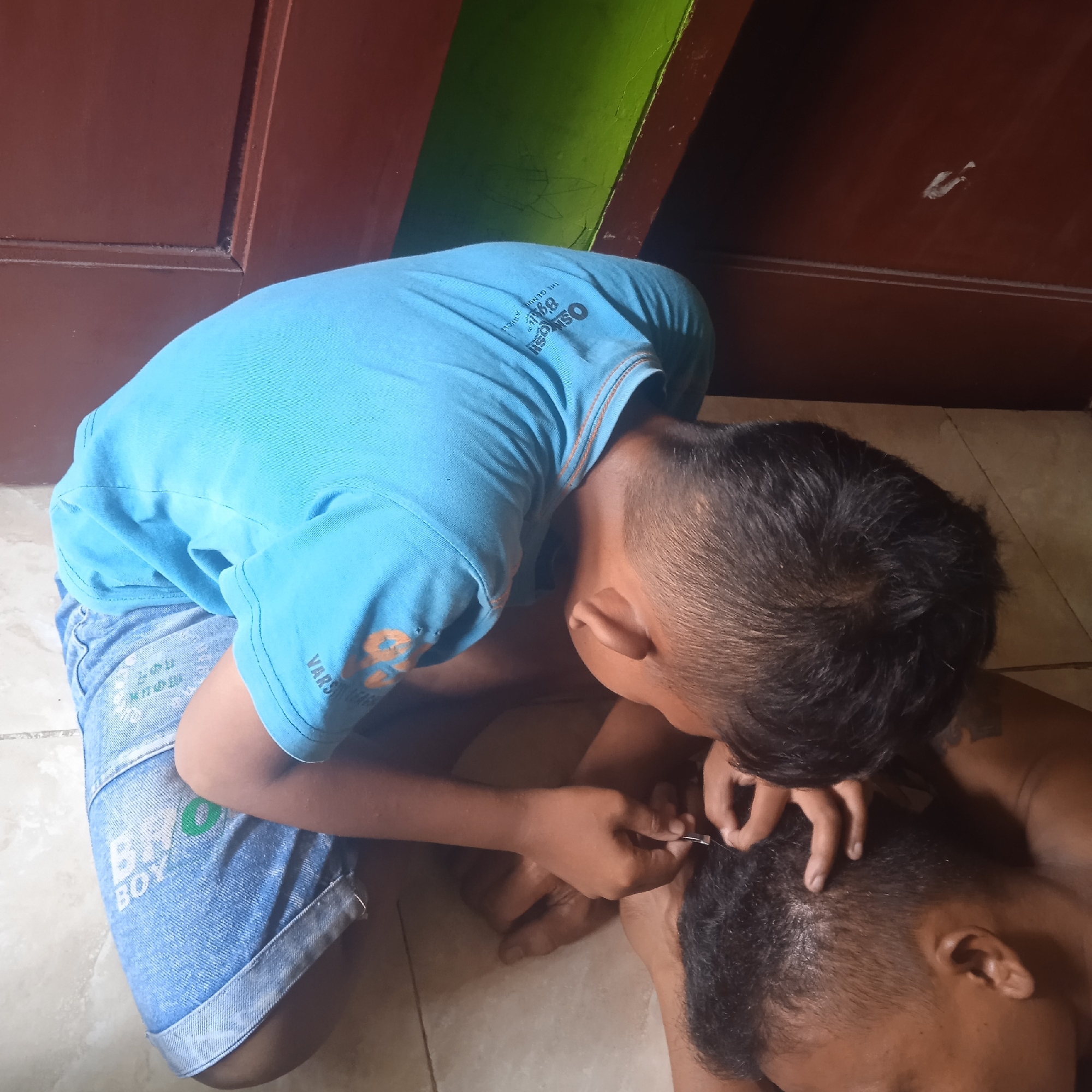 The image shows two young boys in what appears to be a home setting in Situterate, Indonesia. One boy, wearing a blue t-shirt and denim shorts, is leaning over another boy, who is lying down. The boy in the blue t-shirt is holding a small pair of scissors and appears to be giving the other boy a haircut or trimming his hair.
The scene is indoors, with a brown wooden door or wall visible in the background, along with a section of a brightly painted green wall. The floor is tiled. The lighting suggests it is daytime, with natural light possibly coming from a window not visible in the frame.
On the blue t-shirt, there is visible text that appears to be a brand logo, "OshKosh," and possibly other words like "THE GENUINE A/CU." The denim shorts also have some white text, with "BOY" clearly legible. There are also some faint markings or graffiti on the green wall in the background.
The activity depicts a personal grooming moment, likely between siblings or friends. The focus is on the intimate act of cutting hair, with the boy holding the scissors concentrating on his task. The overall atmosphere seems casual and domestic.