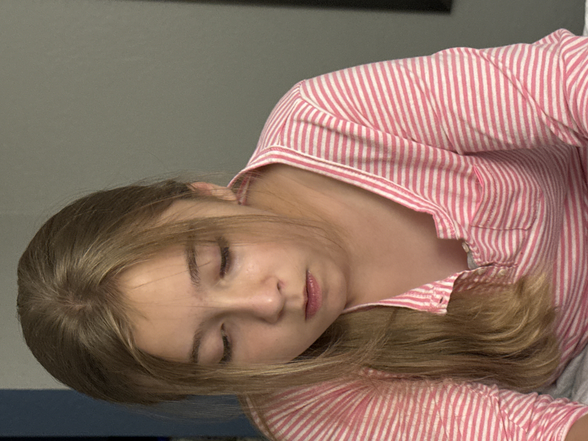 A young girl with blonde hair pulled back is looking down. She is wearing a pink and white striped shirt. The background is a plain gray wall with a blue object to the left. The image appears to be taken indoors, possibly in a home setting, in Myrtle Beach, United States of America.