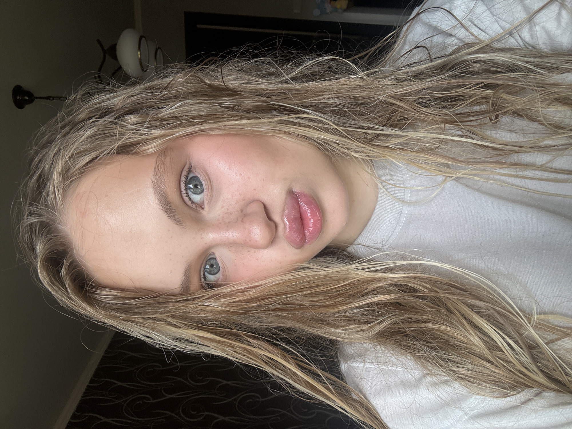The media file is a portrait of a young woman with blonde hair. She is wearing a white t-shirt. Her eyes are open and she is looking at the camera. Her lips are slightly parted and appear to be glossed. She has freckles on her nose and cheeks. Her hair is long and appears to be somewhat damp, with strands falling across her face and shoulders. The background is dark and indistinct, with a hint of a lamp and patterned wallpaper visible on the left side. The lighting suggests it may be indoors, possibly in the evening or a dimly lit room. The overall mood appears casual and relaxed. No visible text is present in the image.