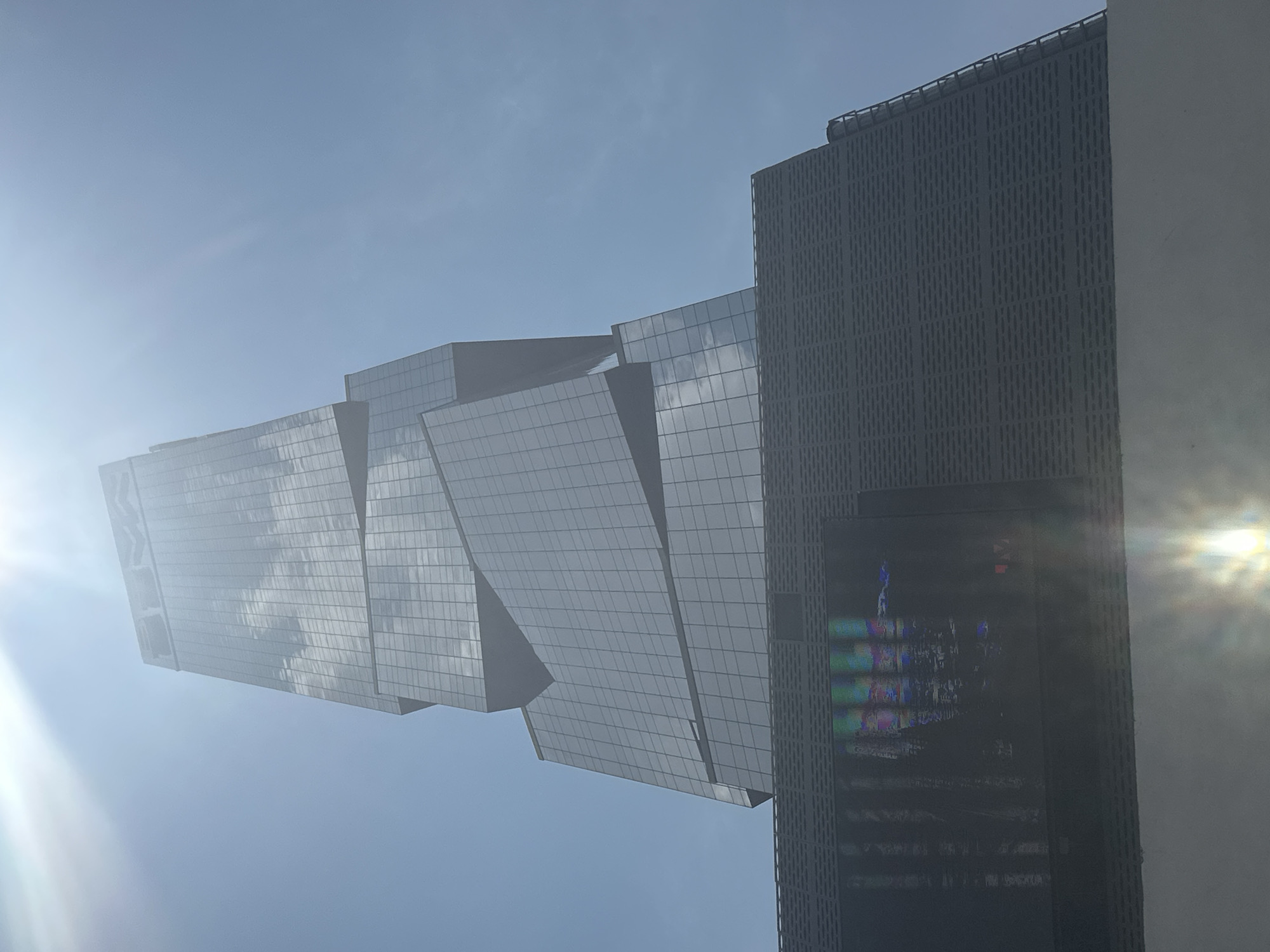The image shows a low-angle view of two modern buildings against a bright blue sky with lens flare. The building on the left has an angular, cantilevered design with a reflective glass facade. The building on the right is taller, with a dark gray exterior and a rectangular shape. It has a large video screen that displays colorful light. The location appears to be an urban area, though San Bernardino Tlaxcalancingo, Mexico is not apparent from the image. No people or specific objects beyond the buildings are visible. The interaction is limited to the structural connection between the two buildings.