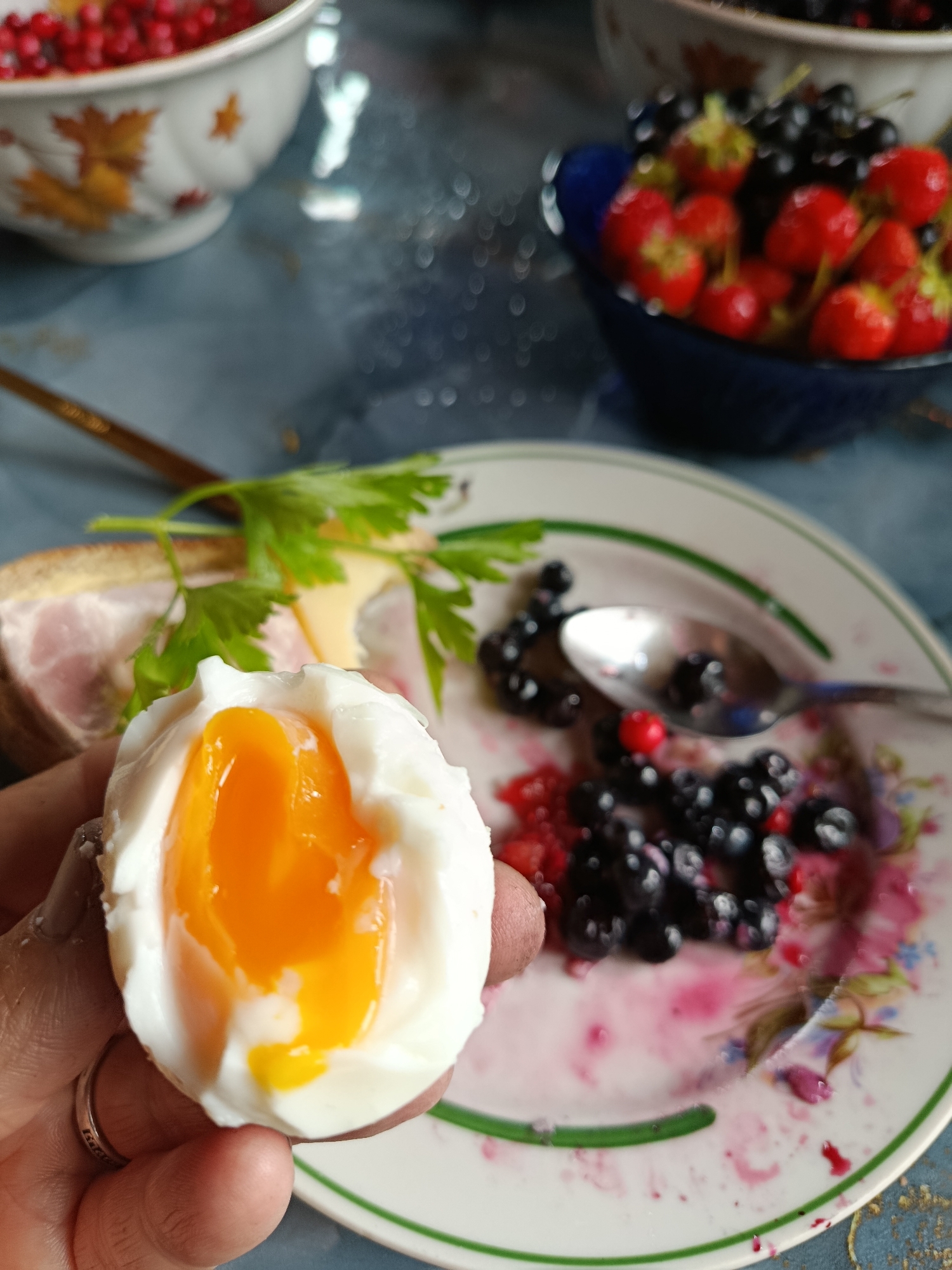 A close-up, overhead shot shows a hand holding a soft-boiled egg with a runny yolk. The egg is positioned in the foreground. Behind it, a plate contains blueberries, raspberries, and a spoon. Further in the background, a bowl of red berries and another bowl of strawberries and blueberries are visible, as well as a sandwich with ham and cheese. The setting appears to be a table with a blue tablecloth. The image quality is somewhat blurry, with a shallow depth of field. The location, Верея, Russia, is mentioned, suggesting a domestic setting.