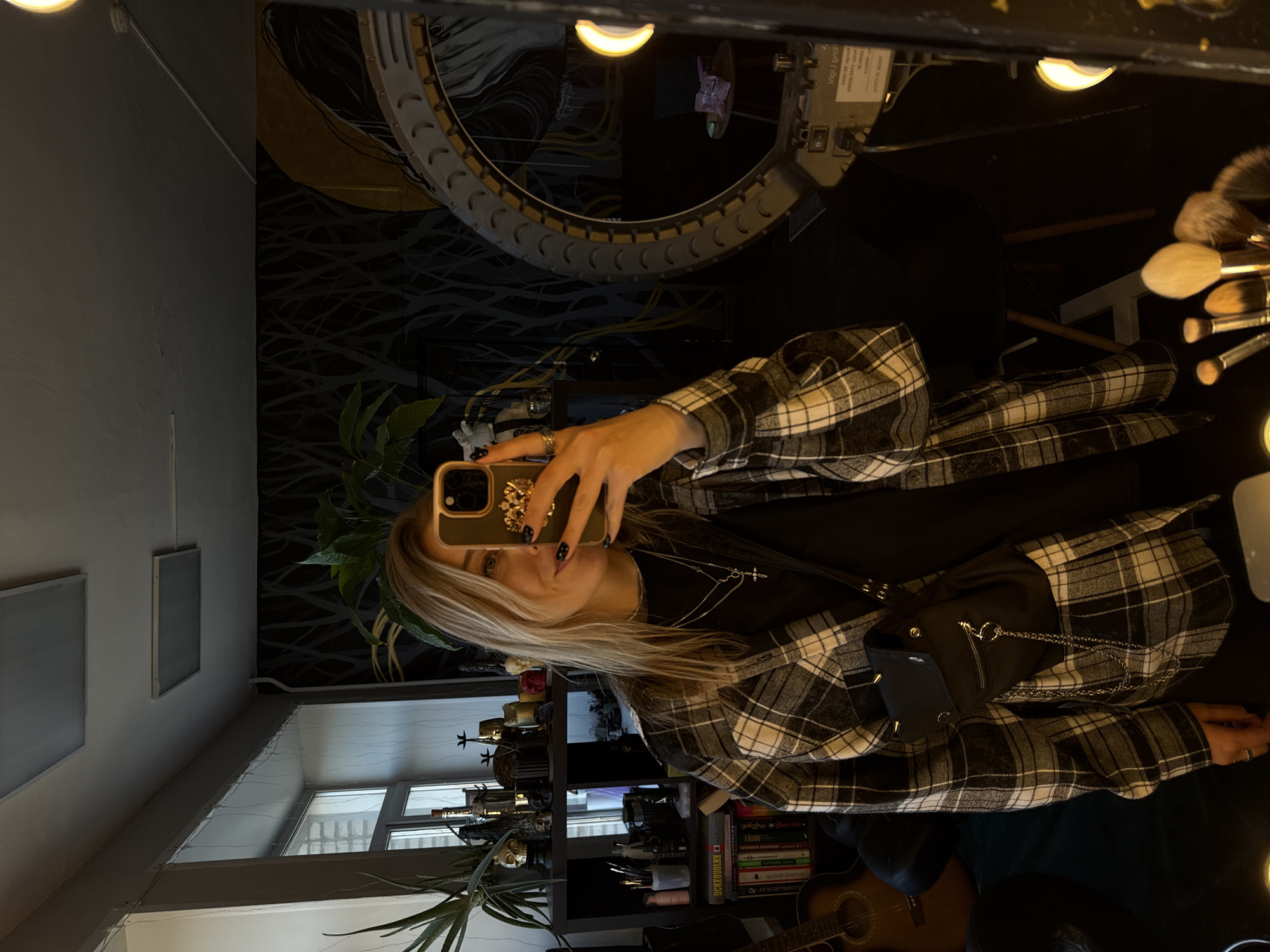 A person is taking a selfie in a mirror. The background shows a room with decorative elements, a ring light, and shelves with objects. A plant is visible near the window. The location may be in Krasnoyarsk, Russia.