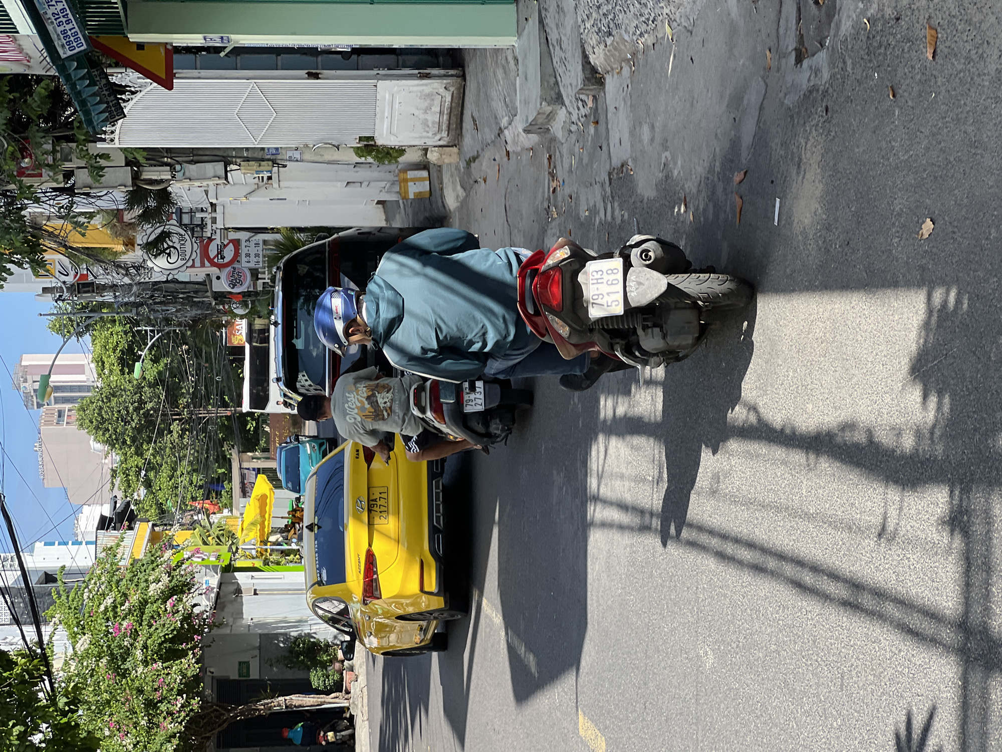 The image shows a street scene in Nha Trang, Vietnam, with a bright yellow car and two motorcycles on a paved road. In the foreground, a person wearing a blue helmet and a teal jacket rides a black motorcycle with license plate "79-H3 5168". Behind them, a yellow Hyundai Accent car with license plate "79A 217.71" is driving. Another motorcycle is partially visible to the left, with a rider in a grey shirt. The street is lined with buildings, trees, and utility poles with many overhead wires. The buildings are a mix of colors including white, yellow, and green, and some have awnings and signs. The sun is casting shadows on the road, indicating it is daytime. The overall impression is of a typical urban street in Vietnam.