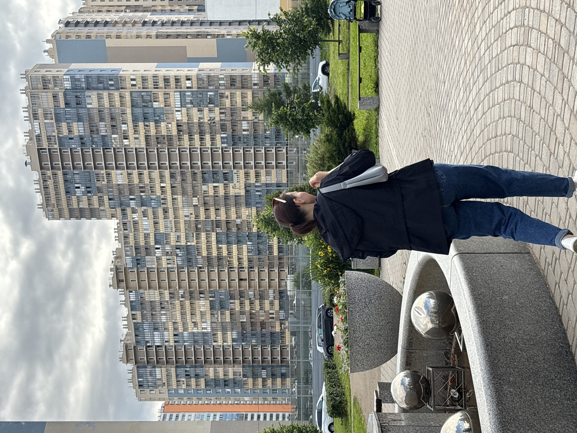 The image depicts a street scene in Saint Petersburg, Russia, likely in a modern residential area. In the foreground, a woman with brown hair pulled back in a ponytail is seen from behind, wearing a black hooded jacket and blue jeans. She carries a light-colored shoulder bag. She is walking on a patterned brick sidewalk next to a stone fountain with metallic spheres. Behind her, a large, multi-story residential building with a facade of varying shades of beige, gray, and blue is prominently featured. The building has numerous windows and balconies. To the left and right of the building are other similar structures. In the middle ground, there is a well-maintained green lawn with trees, benches, and a flower bed in a large, textured planter. Several cars are visible on a road behind the lawn, and a baby stroller is parked on the grass. The sky is overcast.