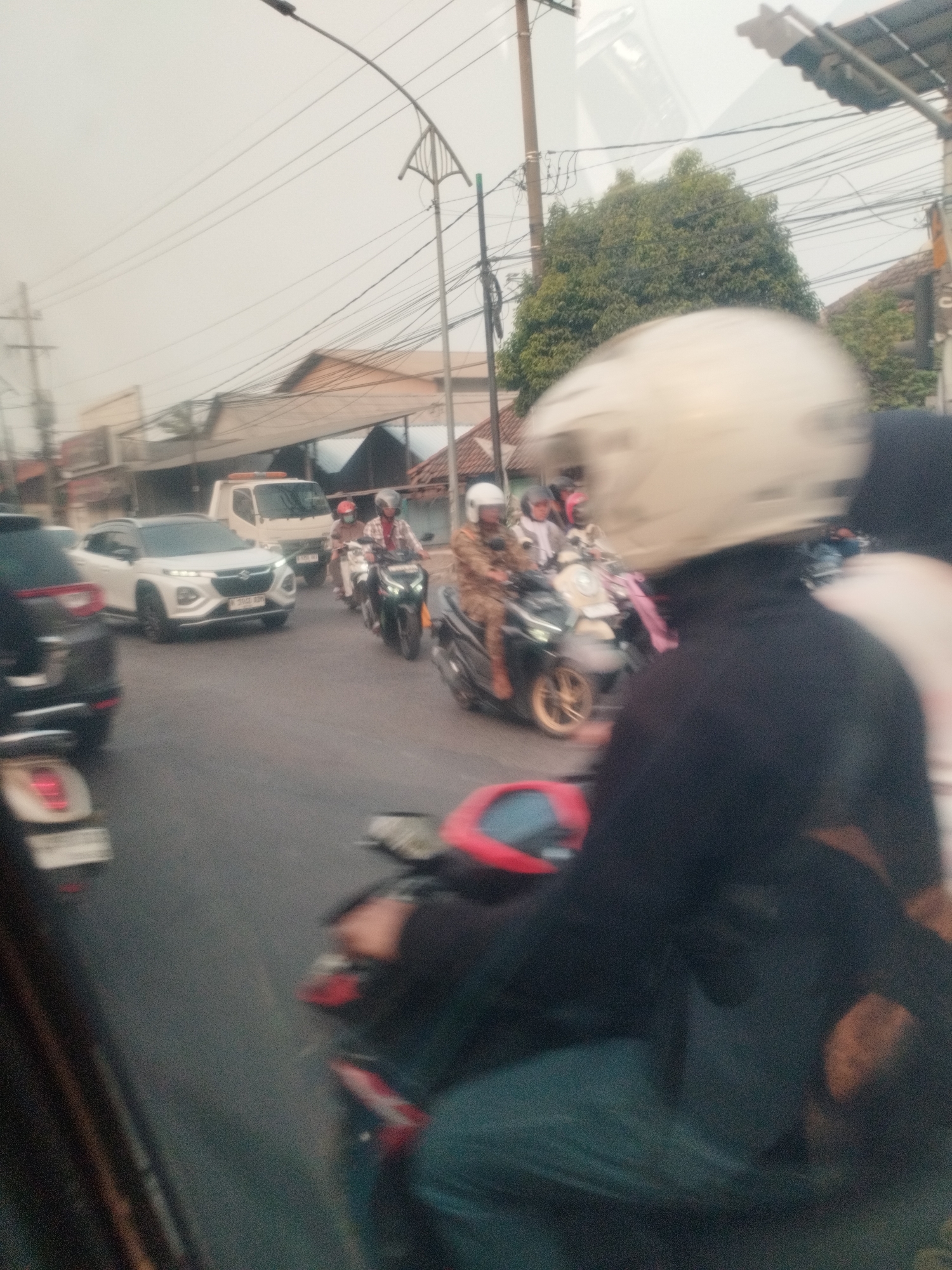 The image captures a street scene in Surabaya, Indonesia, characterized by a busy road with various vehicles. In the foreground, a person wearing a white helmet on a motorcycle is the primary subject, with their form blurred due to motion. Behind them, other motorcycles and a white car are visible, also in motion. The car appears to be a modern SUV with its headlights on. Further in the background, a white truck and buildings lining the street can be seen. Overhead, power lines crisscross the sky, a common sight in urban Indonesian settings. The overall atmosphere suggests a typical traffic situation in a bustling city.