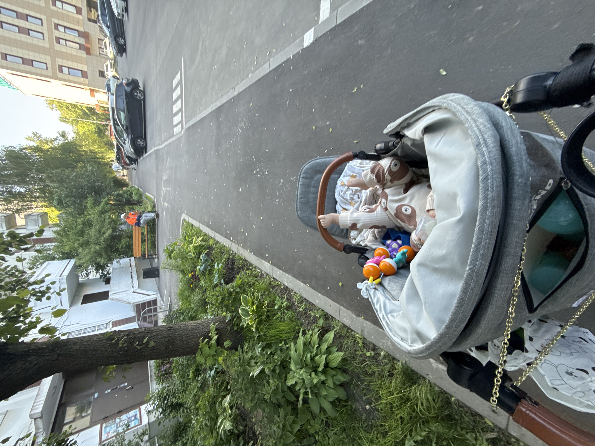 This media file captures an outdoor urban scene during what appears to be a clear, sunny day, likely in Moscow, Russia. The primary subject is a baby resting in a gray stroller, positioned on a paved sidewalk next to a road. The baby is dressed in a light-colored outfit with a brown pattern, possibly featuring bears or koalas, and is partly covered by a white blanket. Several colorful infant toys, resembling rattles, are visible near the baby's feet within the stroller. The stroller itself has a brown-toned handle.
To the left of the stroller, a strip of green foliage, including bushes and a large tree trunk, separates the sidewalk from residential buildings. These multi-story buildings, typical of urban settings, are visible in the background. The paved road features white pedestrian crossing markings and several parked cars. Further down the sidewalk, in the mid-ground, two adults are seen sitting on a wooden bench; one is notably wearing an orange vest. The overall impression is a peaceful moment during a stroll. No discernible text is clearly visible within the image.