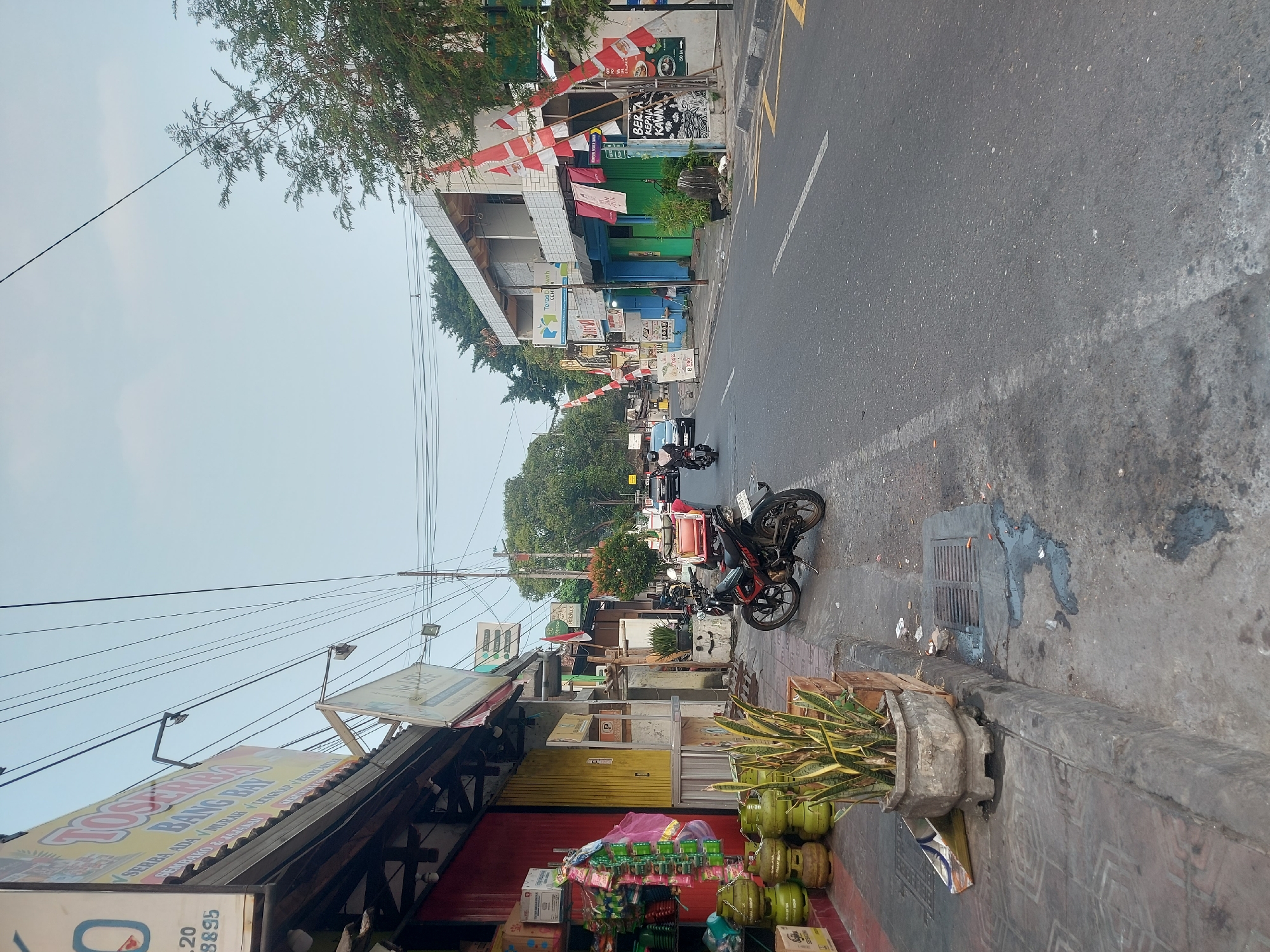 The image depicts a street scene in Yogyakarta, Indonesia, with shops lining the left side of the road and a busy street on the right. On the left, several shops are visible, one with a red facade and shelves displaying goods, including gas canisters and packaged items. Above the shops, large yellow and white signs are affixed, with text in Indonesian, including one that reads "TOSEARBA BANG BAY." A planter with a snake plant stands on the sidewalk in front of one of the shops.
The street itself is paved and appears to be a main thoroughfare. Several vehicles are present, including two motorcycles parked on the side and one motorcycle and a car driving on the road. Red and white flags, likely Indonesian flags, are prominently displayed on poles along the sidewalk on the right side of the street, suggesting a national holiday or celebration. The buildings on the right side of the street vary in style, with some appearing more modern and others older. Trees and greenery are visible in the background, adding to the urban but natural landscape. The sky is overcast, indicating a cloudy or hazy day. The overall impression is of a lively, commercial street in a Southeast Asian city.