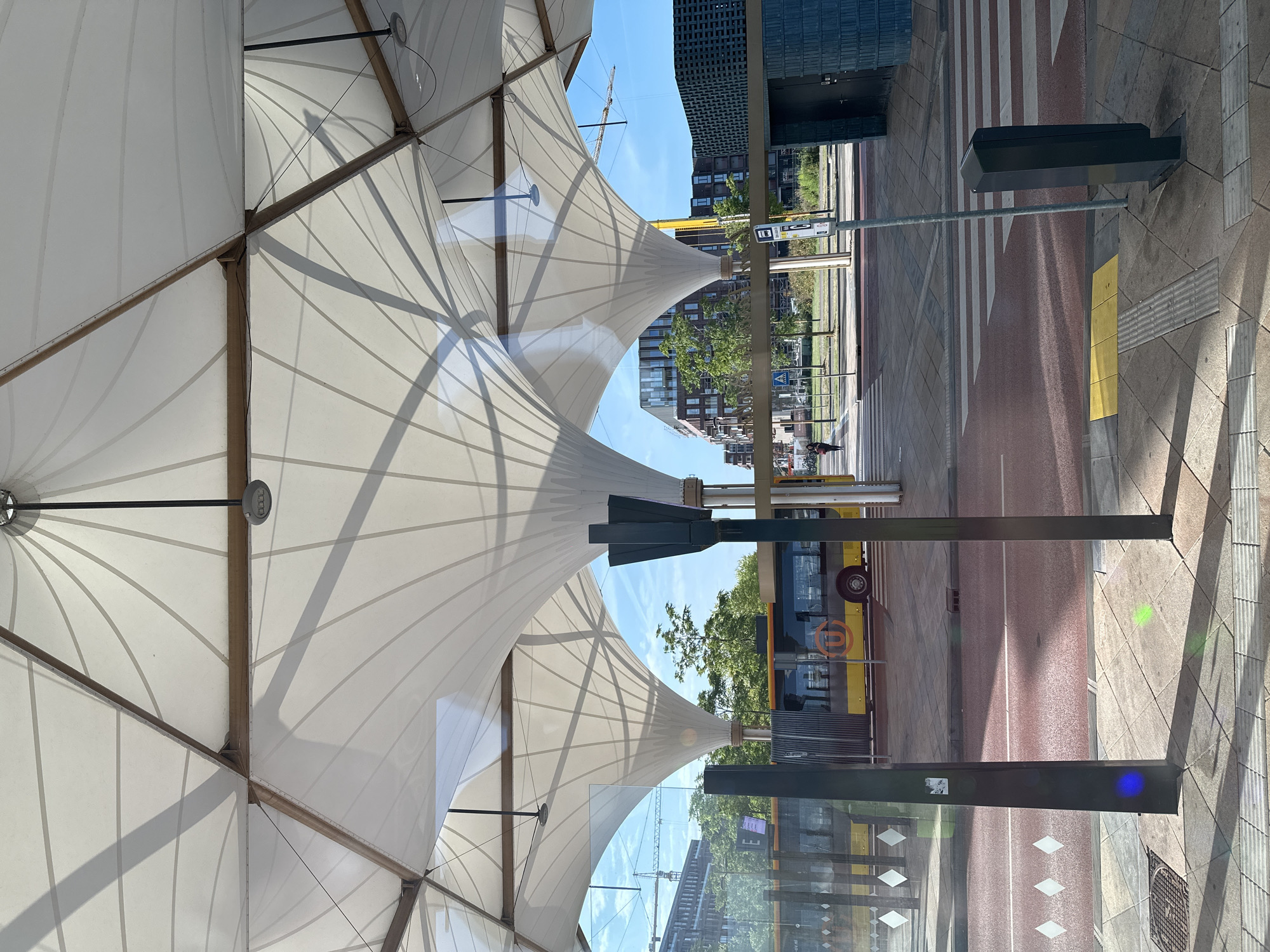 This is a photograph taken from underneath a modern, white, tensile fabric structure at a bus station. The structure has a hexagonal grid pattern formed by wooden beams and white fabric panels, creating a series of undulating curves. Lights are visible attached to the beams. Through the openings of the structure, a sunny day with a blue sky is visible.
In the background, a yellow and grey bus is stopped at a bus stop. The bus has the "U" logo of "U-OV" (Utrecht Public Transport) visible on its side. The bus stop area has a red-colored road surface with white pedestrian crossings and yellow tactile paving. Beyond the bus stop, there are modern buildings with brick facades and green trees. Construction cranes are also visible in the distance, suggesting ongoing development in the area.
A person is walking across the pedestrian crossing in the mid-ground. There is a bus stop sign and a dark gray bollard near the pedestrian crossing. Reflections of blue and green light are visible on the foreground pavement. The overall scene depicts a functional urban public transport hub on a clear day. This location is likely the Utrecht Central Station bus terminal, given the U-OV bus and the modern infrastructure consistent with major Dutch transport hubs.
