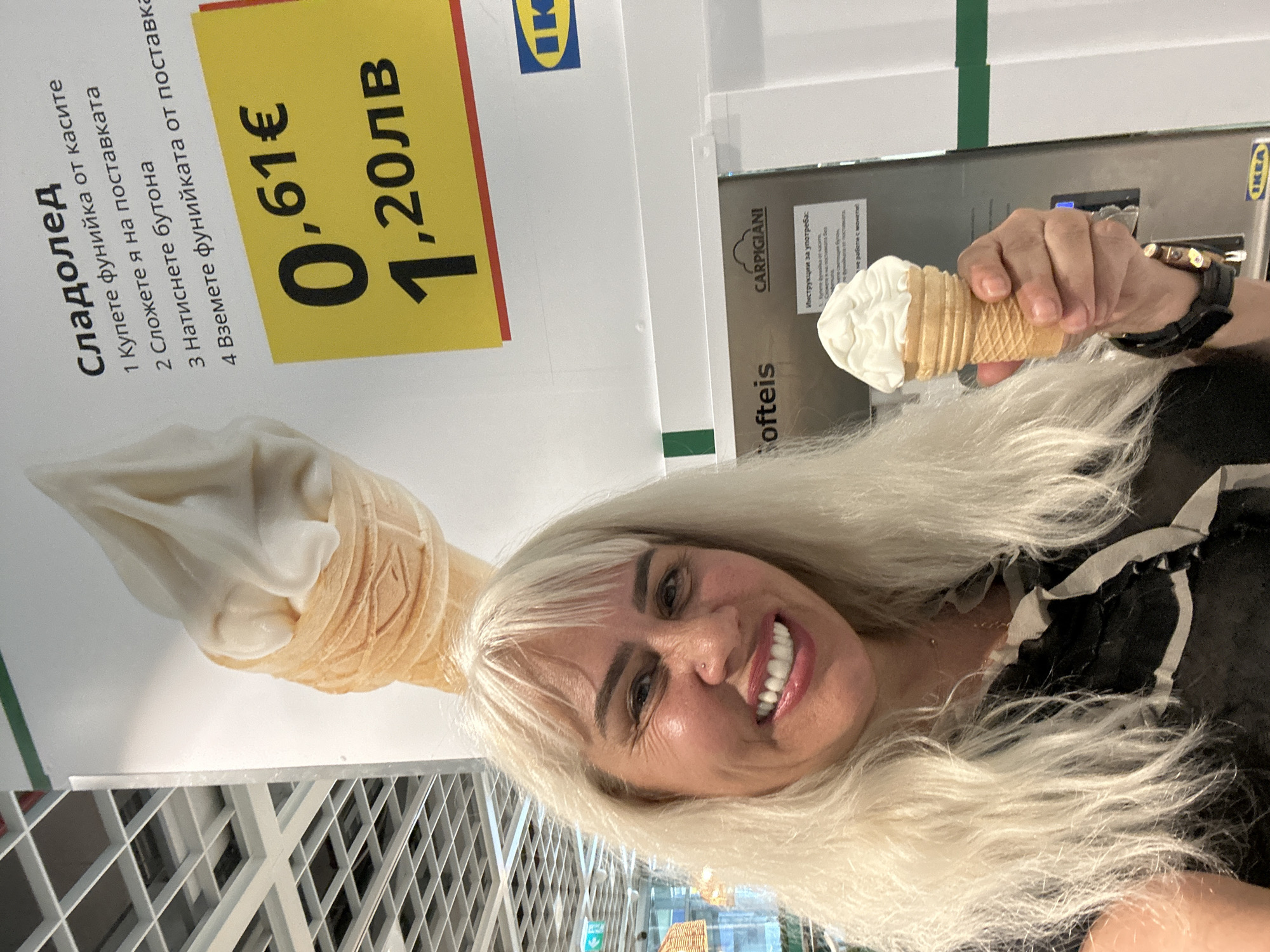 Here is a description of the image:
A blonde woman is holding an IKEA soft-serve ice cream cone in her right hand, smiling and looking directly at the camera. She is wearing a black wristwatch on her left wrist and has a visible nose ring. Behind her is an IKEA store, specifically a large advertisement for the same soft-serve ice cream cone she is holding. The poster includes text in Bulgarian: "Сладолед" (ice cream), followed by four instructions on how to obtain the ice cream. It also shows the price in both euros (€0.61) and Bulgarian lev (1.20лв). Next to the poster is a Carpigiani soft-serve ice cream machine. The background has a slightly blurry view of the interior structure of the IKEA store. The location is likely within the IKEA store in Sofia, Bulgaria.