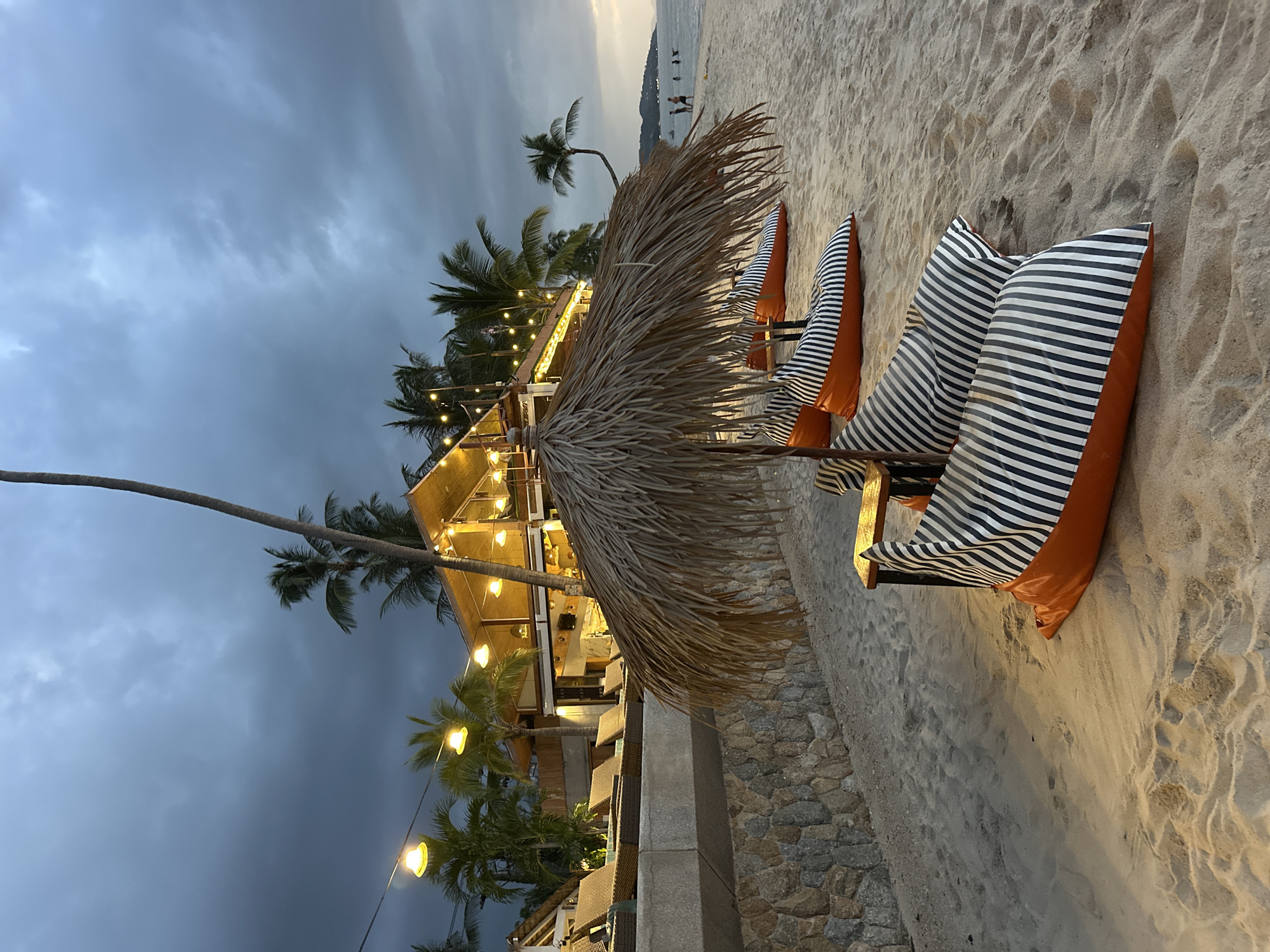 The image depicts a beach scene at dusk in Ko Samui District, Thailand. In the foreground, several beanbag chairs with orange and black-and-white striped covers are arranged on the sandy beach. A large thatched umbrella shades some of the seating. Behind the beanbags, a wall made of rough stone separates the beach from a raised area with lounge chairs and a building structure with a sloped, thatched roof. This building is illuminated by string lights, suggesting it is a restaurant or bar. Palm trees are visible, adding to the tropical atmosphere. The sky is overcast with dramatic clouds, and the sea is calm with a few distant figures visible in the water and on the shore. The overall impression is one of a tranquil evening setting at a beachside establishment.