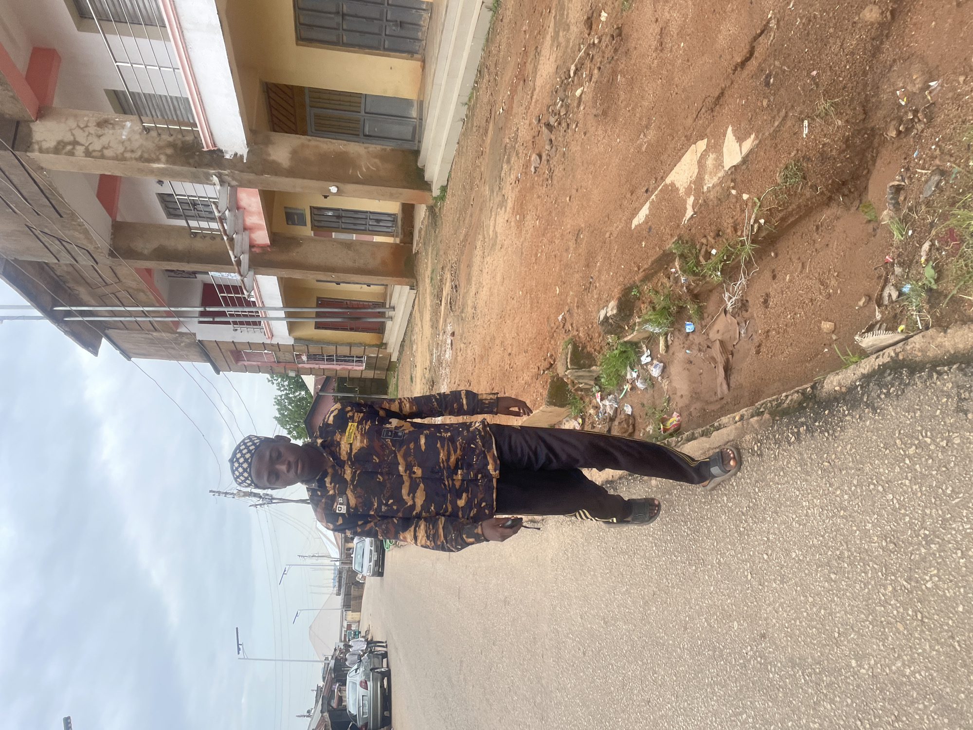 In Jos, Nigeria, a young Black man is walking down the side of a paved road. He is wearing a camouflage patterned long-sleeved shirt, black pants with a yellow stripe on the side, and black sandals. He also wears a patterned skull cap. He is walking towards the camera and holding a dark object in his left hand. To his right, a muddy ditch runs alongside the road, littered with trash and some greenery. Behind the ditch, there is a two-story building with a yellow and pink facade, featuring multiple windows and balconies. The sky is overcast. In the background, other buildings and vehicles are visible, suggesting a populated urban area.