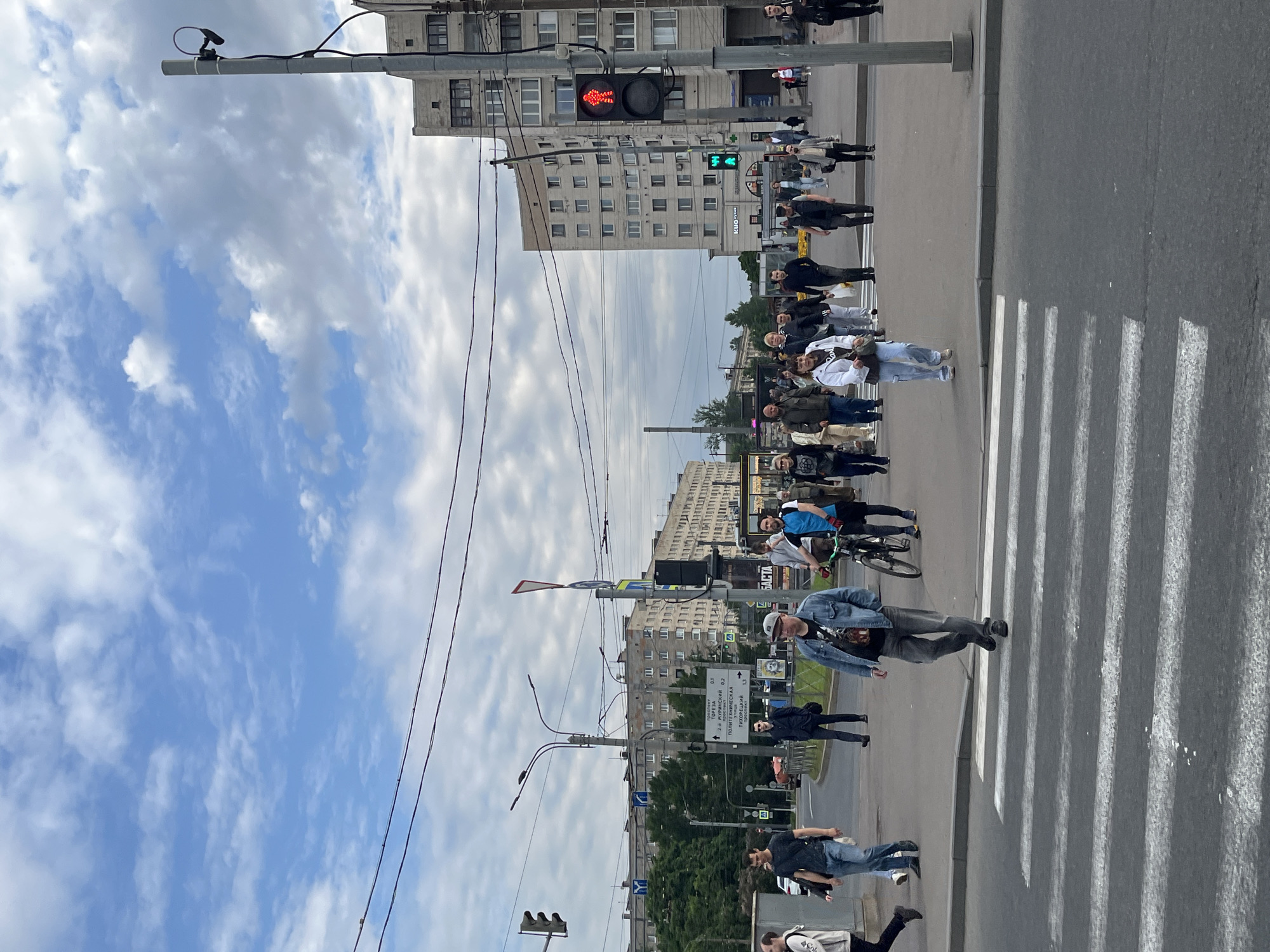 This urban scene captures a busy street intersection in Saint Petersburg, Russia, under a partly cloudy sky during daylight hours. The image is filled with pedestrians, buildings, and typical city infrastructure.
In the foreground, a wide pedestrian crosswalk (zebra crossing) spans the asphalt road. Numerous people are gathered on the sidewalk to the right of the crosswalk, waiting for the signal to change, while a man in a denim jacket and jeans is actively crossing the street. Further back, another person is on a bicycle. The pedestrians are dressed in casual, modern clothing.
Prominently featured are traffic signals; a pedestrian traffic light clearly displays a red "don't walk" figure. Overhead power lines and street lighting poles crisscross the sky, with various cameras or sensors attached to the poles. Multi-story residential or commercial buildings line both sides of the street, featuring numerous windows and suggesting a dense urban environment.
A white directional sign with Cyrillic text is visible on a street pole. The text indicates street names and distances, including "ТОРЕЗА" (Toreza), "ПОЛИТЕХНИЧЕСКАЯ" (Politekhnicheskaya), "2-Й МУРИНСКИЙ" (2nd Murinsky), "КУРЧАТОВСКИЙ" (Kurchatovsky), and "ТИХОРЕЦКИЙ" (Tikhoretsky), along with numerical distances like 0.1, 0.2, 0.6, and 1.3. On a building in the background, partial text "KUMO" and "OPHT" can be seen, likely part of a shop or clinic sign. The overall mood is one of everyday urban activity, with people going about their routines.
