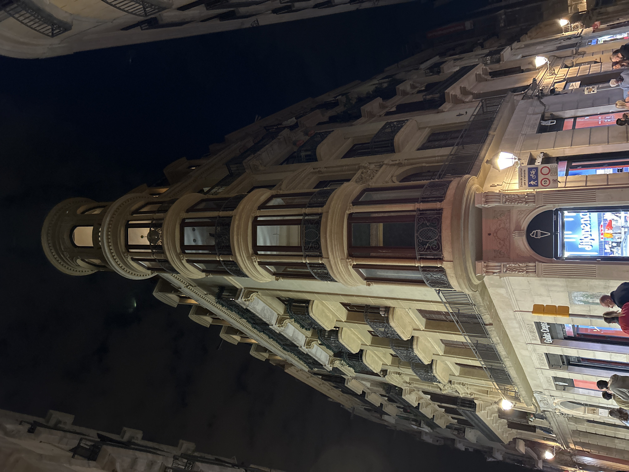 Captured at night, the image showcases a building facade in Barcelona, Spain. The architectural style is ornate, with multiple floors, balconies, and decorative elements. The building is illuminated by streetlights, highlighting its details. People are present on the sidewalk in front of the building. Shopfronts are visible at street level. The overall environment is urban.