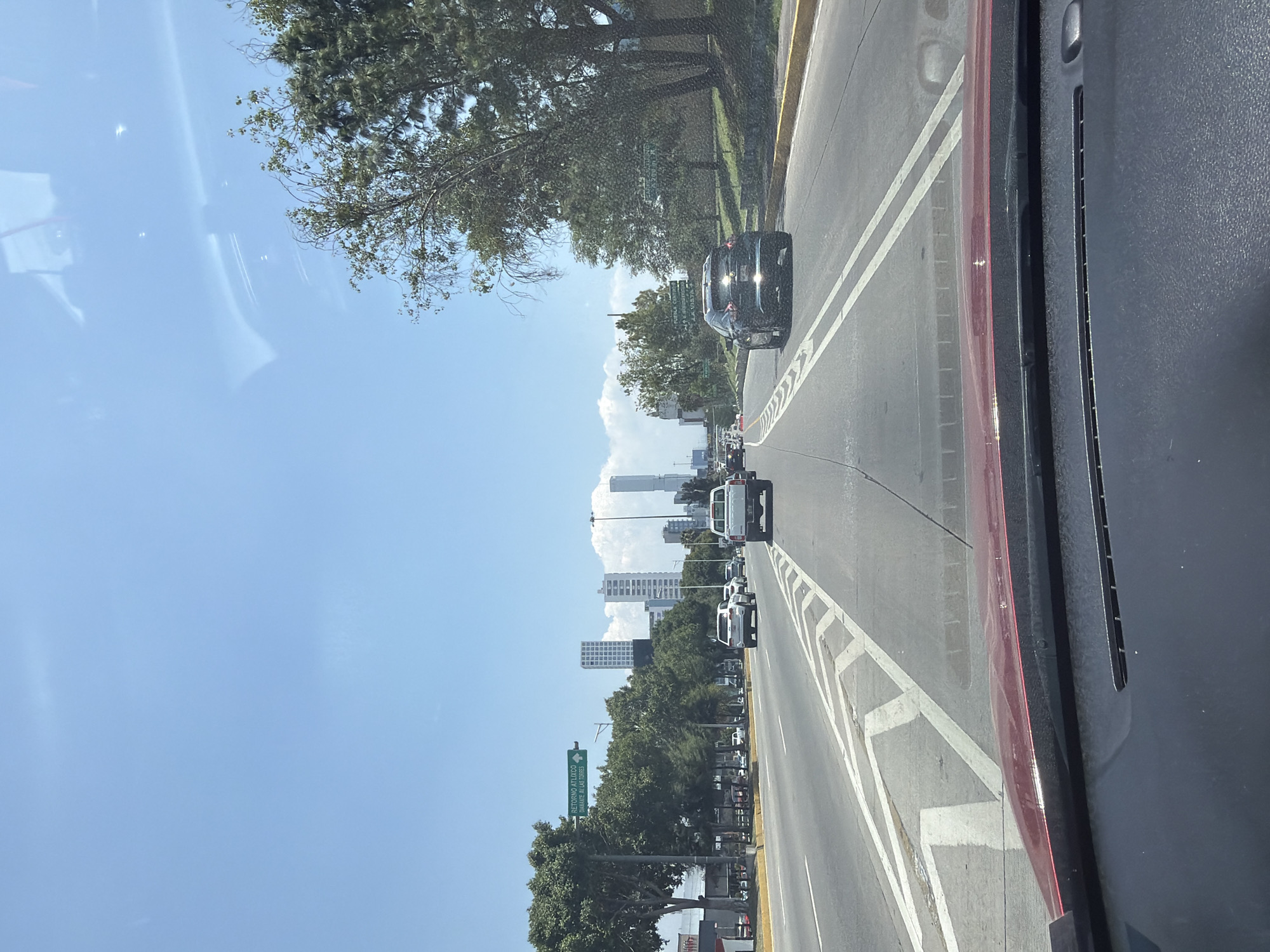 The image, taken from inside a vehicle looking through the windshield, depicts a multi-lane road stretching into the distance under a clear, sunny sky. The road features white lane markings, hatched areas, and a yellow line along the left shoulder. Several vehicles are visible, including a dark-colored car in the right foreground lane and a line of white vehicles (vans or SUVs) further down the road.
Lush green trees line both sides of the thoroughfare, with a dense row along the left. In the background, a modern city skyline with several tall buildings rises against the horizon, under a blue sky adorned with scattered white clouds. The bright illumination suggests it is daytime, likely mid-morning or early afternoon.
The right side of the image shows a portion of the vehicle's dark dashboard and a red trim detail. Reflections of the vehicle's interior are visible on the windshield, particularly in the upper left. A green road sign is partially visible on the left side of the road, through the trees. The text on the sign clearly reads "RETORNO ATLIXCO" and below it "AV. LA PAZ - AV. LAS TORRES," providing a strong location cue indicating the scene is in Mexico, consistent with San Bernardino Tlaxcalancingo. The overall scene captures travel towards an urban area on a clear, pleasant day.