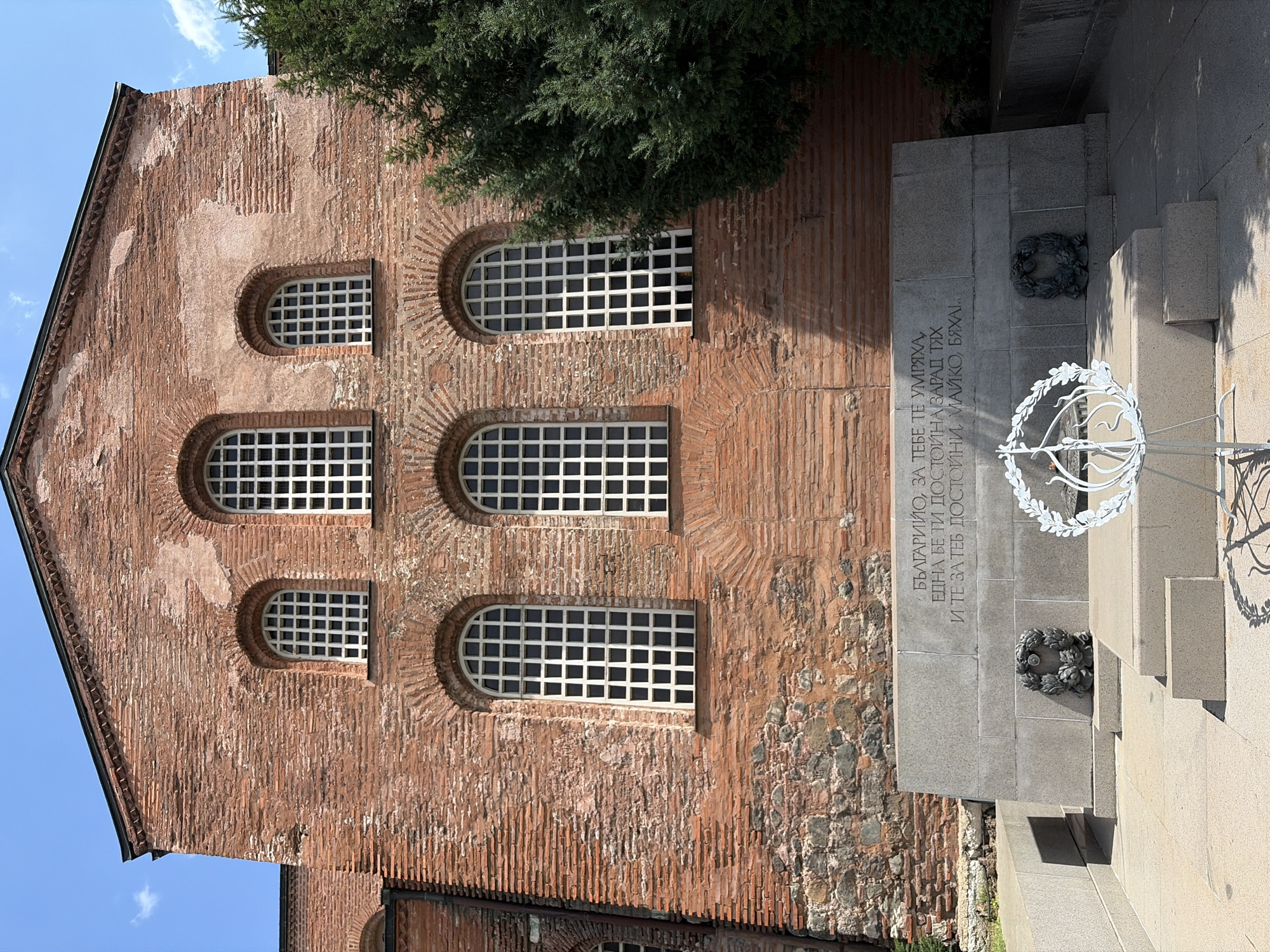 The image shows a monument in front of the Church of St. Sophia in Sofia, Bulgaria. The church is a large brick building with a dark roof and arched windows with white grates. A large stone monument with an inscription in Cyrillic is in the foreground. There are two wreaths on either side of the monument. A white metal wreath stand with a candle is in front of the monument. A large evergreen tree is to the right of the church. The sky is blue with some clouds.