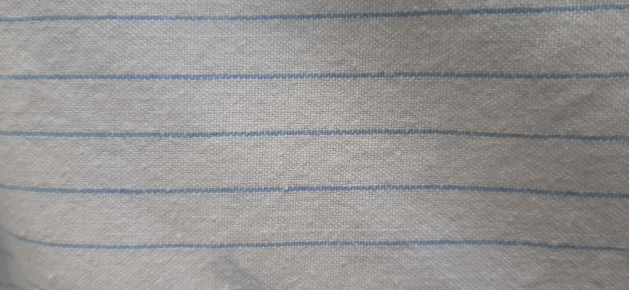 The image is a close-up, top-down view of a light-colored fabric with thin, horizontal blue stripes. The fabric appears to be woven, with a slightly rough texture visible. The stripes are evenly spaced and run across the entire frame. There are no people, objects, or environmental details visible that would suggest a specific location within Yessentukskaya, Russia, or any other place. The image solely depicts the texture and pattern of the fabric.