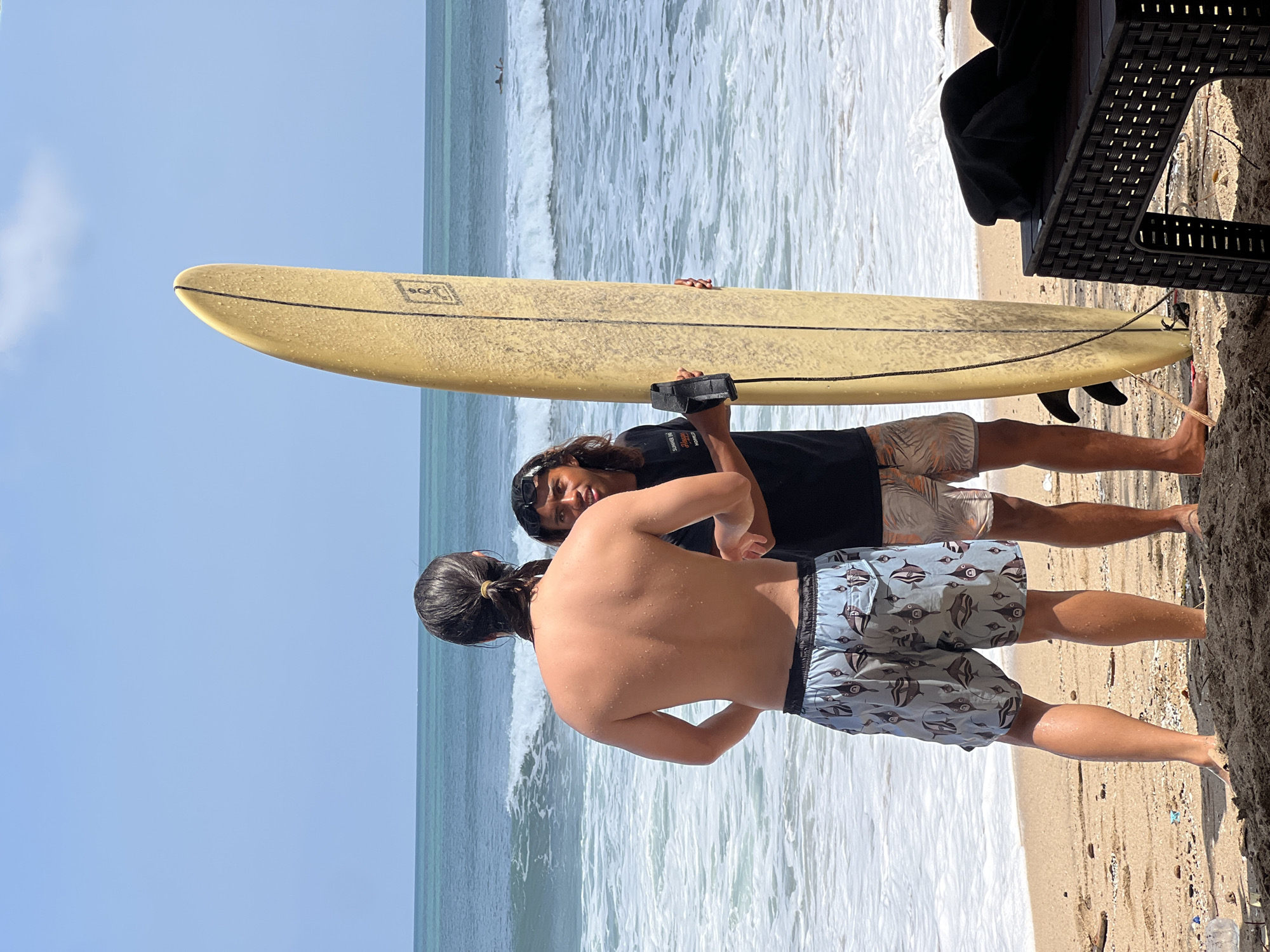 Two men are standing on a sandy beach in Kuta, Indonesia, with the ocean and waves in the background. The man on the left, viewed from behind, has long dark hair tied back and is wearing patterned swim trunks. The man on the right, facing the camera, has long dark hair and is wearing a black t-shirt and patterned swim trunks. He is holding a yellow surfboard upright. The ocean is blue with white waves, and the sky is clear and blue. There are a few pieces of debris on the sand.