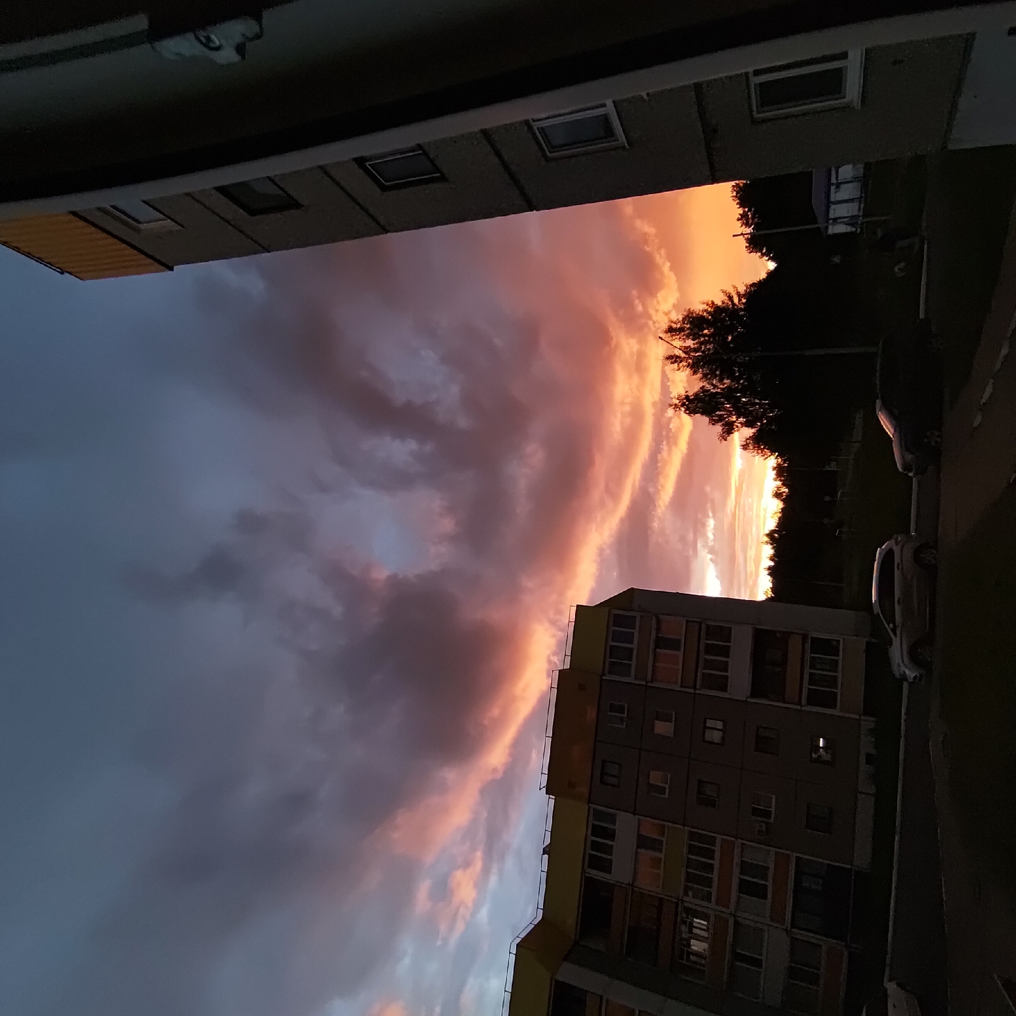 This is a photograph taken from a residential building in Markovsky, Russia, during sunset. The sky is filled with dramatic clouds, illuminated by the setting sun with vibrant hues of orange, pink, and purple, contrasting with the darkening blue sky. In the foreground and mid-ground, the sides of two multi-story apartment buildings are visible. The building on the left has a yellow trim along its upper edge and features multiple balconies and windows. The building on the right is darker and partially obscures the view. In the lower portion of the image, two cars are parked on a paved area in front of the buildings. A line of trees forms a dark silhouette against the illuminated sky in the background, and a streetlamp is visible among them. The overall scene captures a typical urban residential environment under a striking sunset sky.