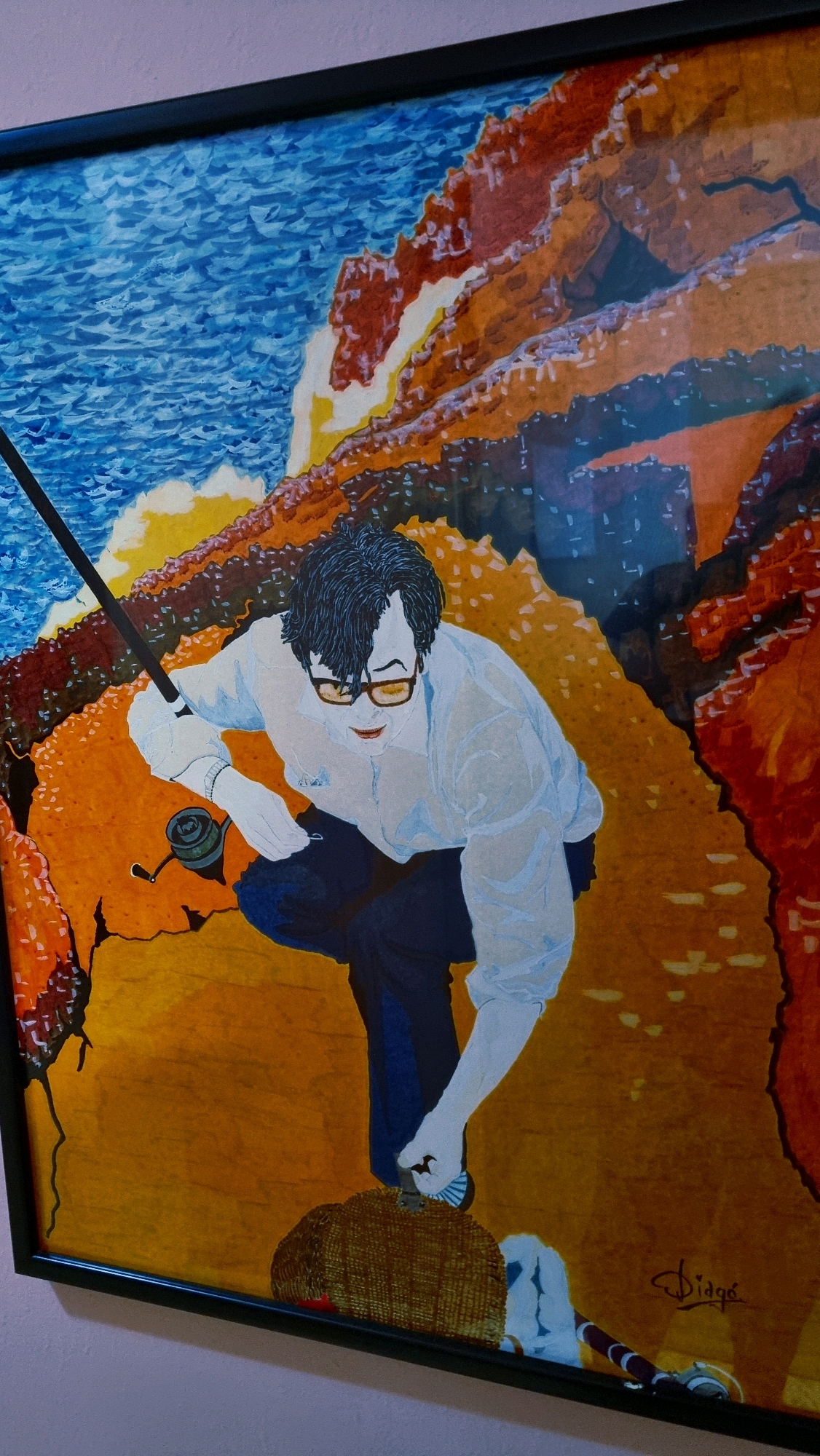 This is a framed painting depicting a man fishing on a rocky coast. The man, with dark hair and glasses, is wearing a light-colored shirt and dark pants. He is holding a fishing rod and appears to be kneeling. The background shows a body of water with waves and reddish-orange cliffs. The signature "Diogo" is visible in the bottom right corner of the painting. The painting is hung on a wall with a light pinkish-purple hue. The location is the municipality of Sant Joan Les Fonts, Spain, a region known for its volcanic landscapes and rivers, which could be the inspiration for the rocky terrain and water depicted in the artwork.