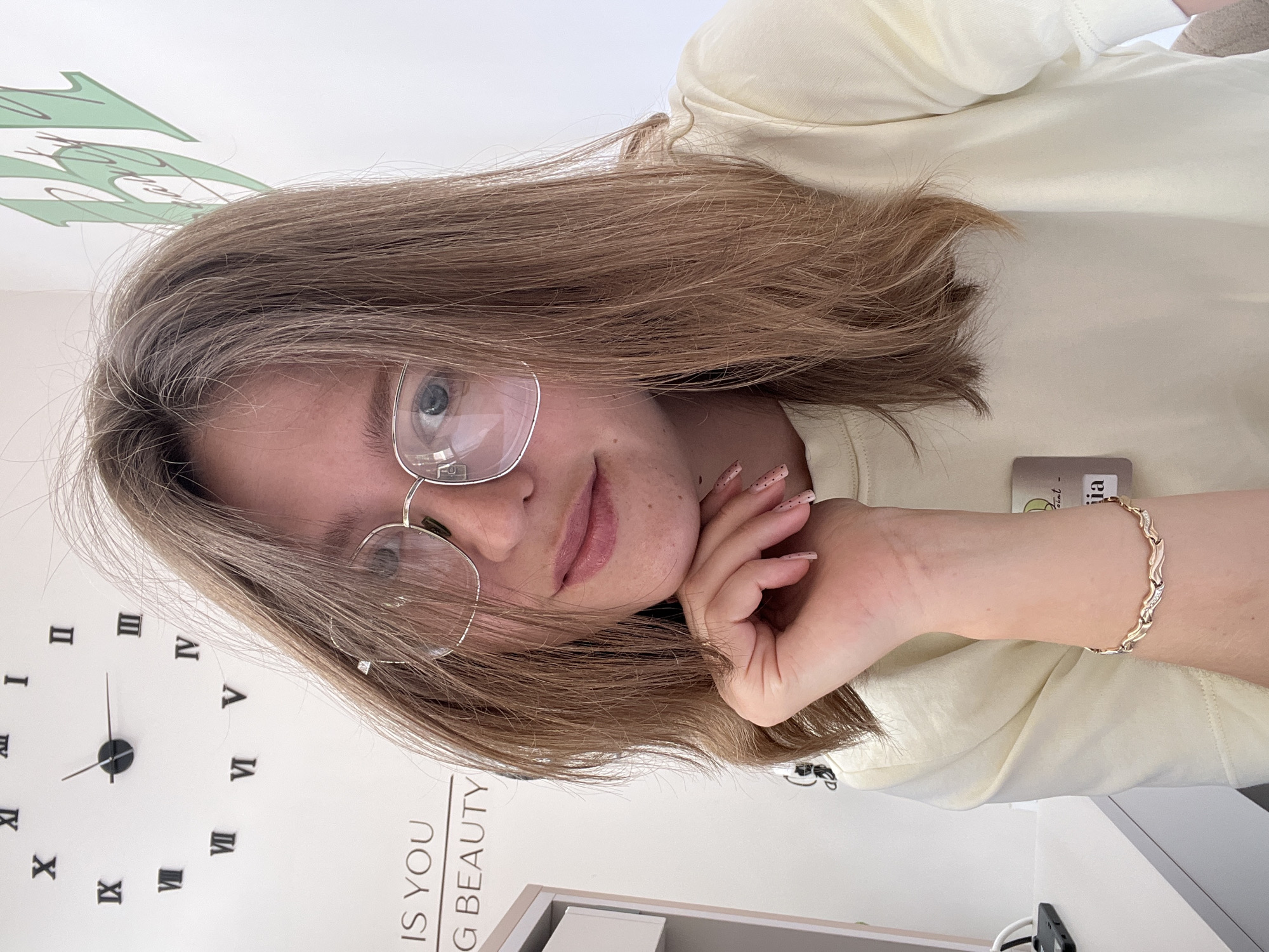 A young woman is depicted in a medium close-up, photographed from a slightly elevated perspective. The image is rotated 90 degrees clockwise, showing her reclining or lying down with her head resting on her right hand. She has long, light brown hair, and wears large, clear-framed eyeglasses that partially obscure her light-colored eyes. Her expression is gentle, with a hint of a smile. Her fingernails are painted with a light, speckled design, and a gold-toned bracelet with a wavy pattern is visible on her right wrist. She is dressed in a plain, light cream or pale yellow t-shirt.
The background consists of a white wall. To her left, a large, modern wall clock with black Roman numerals and hands indicates approximately 10:10. Below the clock, partial text on the wall reads "IS YOU G BEAUTY". To her right, a green, stylized graphic or logo is visible on the wall. A small card or tag with incomplete text, possibly "int - ia", is near her right arm. The setting appears to be indoors, likely a casual environment such as a home or salon in Podgorica, Montenegro.