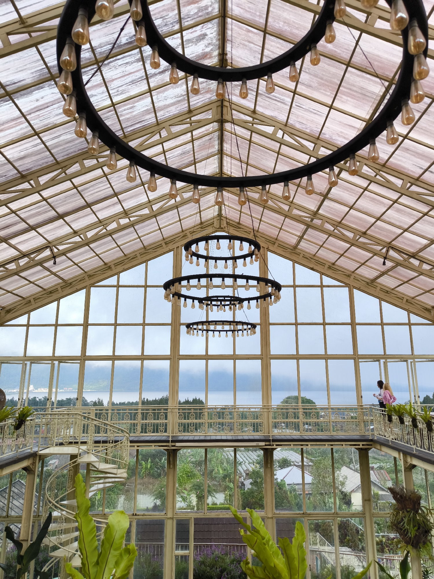 The image shows the interior of a building, likely a greenhouse or glasshouse, in Candikuning, Indonesia. The structure features a high, peaked roof constructed of glass panes held together by a grid of metal supports. Three large, circular chandeliers hang from the ceiling, each adorned with numerous light bulbs. A mezzanine level with a decorative railing runs along one side of the building, and a spiral staircase leads to it. Two people are standing on the mezzanine, observing the outdoor scenery, which includes a body of water, trees, and buildings. Lush greenery is visible both inside the building and through the windows.