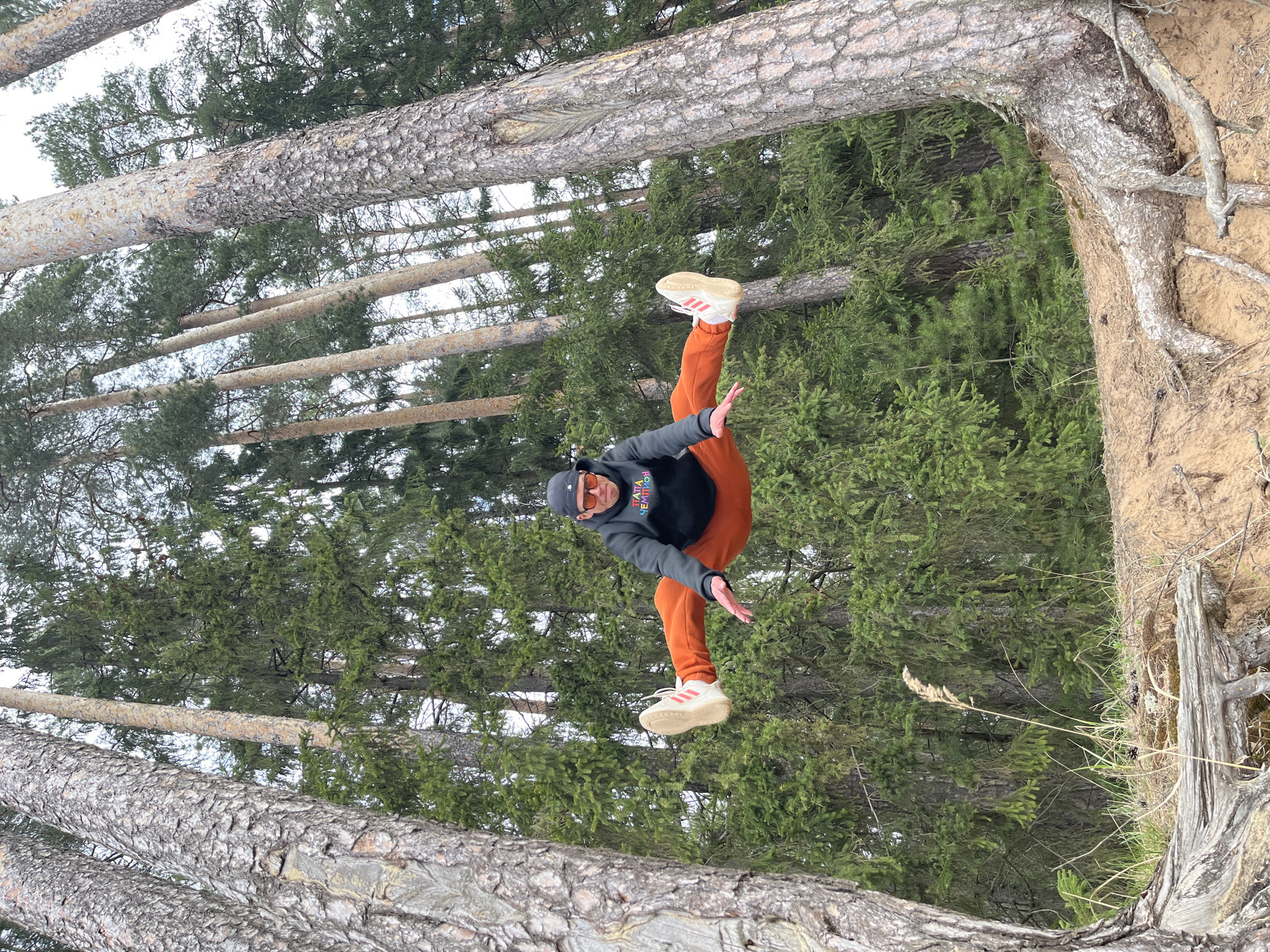 A man in orange pants, a black hoodie, a black baseball cap, and sunglasses jumps in a forest in Chaikovsky, Russia. The man is in mid-air with his legs extended and arms outstretched. Trees with light-colored bark surround him. The ground is covered in green foliage and brown dirt. Some trees appear to be lying on their side, one with exposed roots.