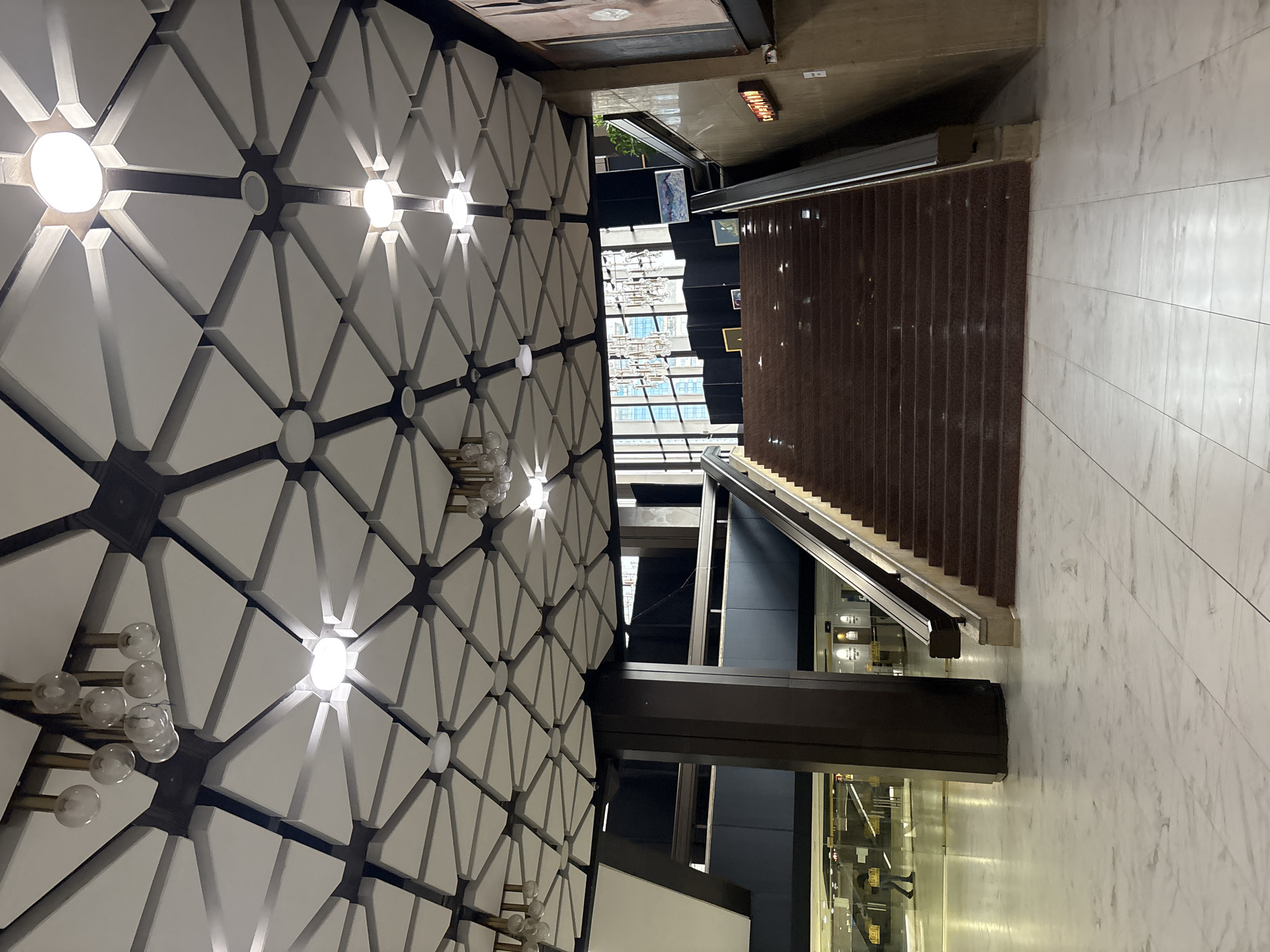 A slightly rotated perspective shows the interior of an airport in Sofia, Bulgaria. A decorative ceiling with a complex geometric pattern of white triangles outlined in dark material and circular lights is visible. An escalator leads up from the ground floor toward a dark brown staircase. Behind the escalators, a glass facade provides a view of a building and an atrium with what appears to be artwork. People are visible within glass-walled corridors below. The floor is composed of marbled tiles.