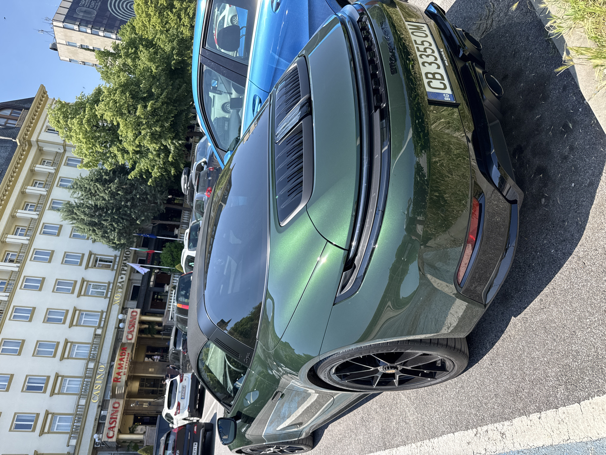 The image depicts a green Porsche parked on a street, adjacent to a building with "Casino Ramada" and "ХОТЕЛ ТРИМОНТИУМ" signage. The location is likely the city of Plovdiv, Bulgaria, based on the presence of the Bulgarian license plate "CB 3355 OM" and the Cyrillic text on the hotel signage. Several other vehicles are parked nearby. The weather appears sunny, with clear skies. The image is taken from a low angle, focusing on the rear of the car.