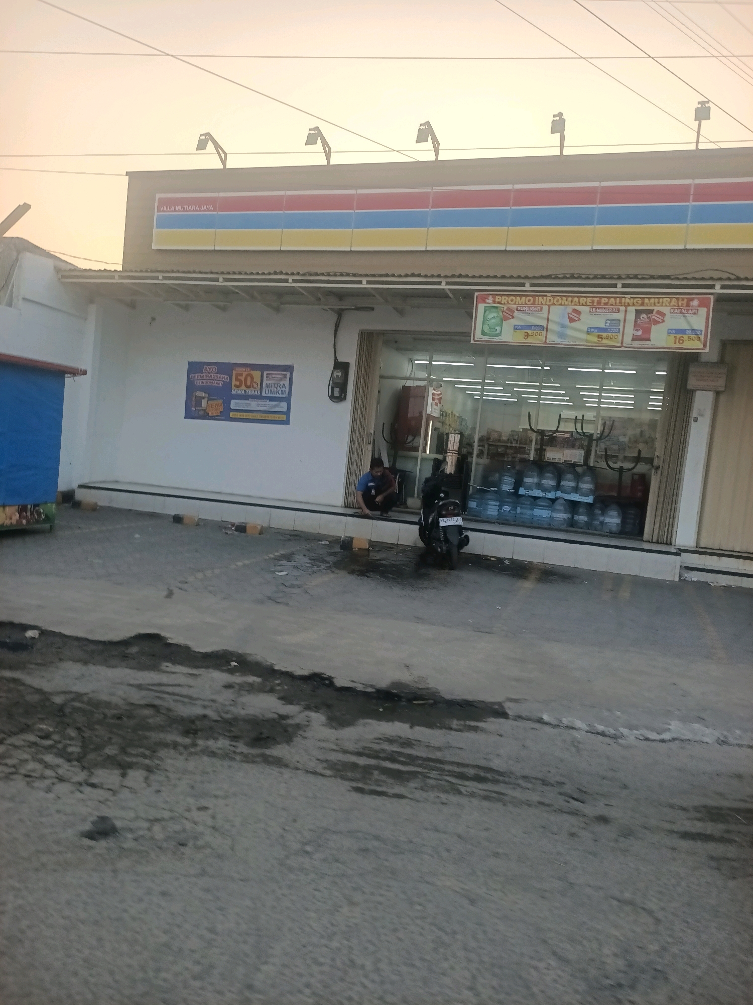 A medium shot captures the facade of a commercial building identified as "VILLA MUTIARA JAYA" above a sign with the Indomaret logo. The building has a light-colored exterior with a covered walkway supported by beams. A banner above the entrance advertises "PROMO INDOMARET PALING MURAH" with images and prices for Sunlight, Le Minerale, and Kapal Api. The storefront features large glass windows revealing shelves stocked with bottled water and other products. Two individuals are visible; one sits on the ground near the entrance, and another, wearing a blue shirt, is seated on the steps. A motorcycle is parked in front of the entrance. The foreground shows a paved area with scattered debris and puddles, suggesting recent rain. The sky is a hazy orange, indicating either dawn or dusk. The scene is situated in the city of Wanajaya, Indonesia.