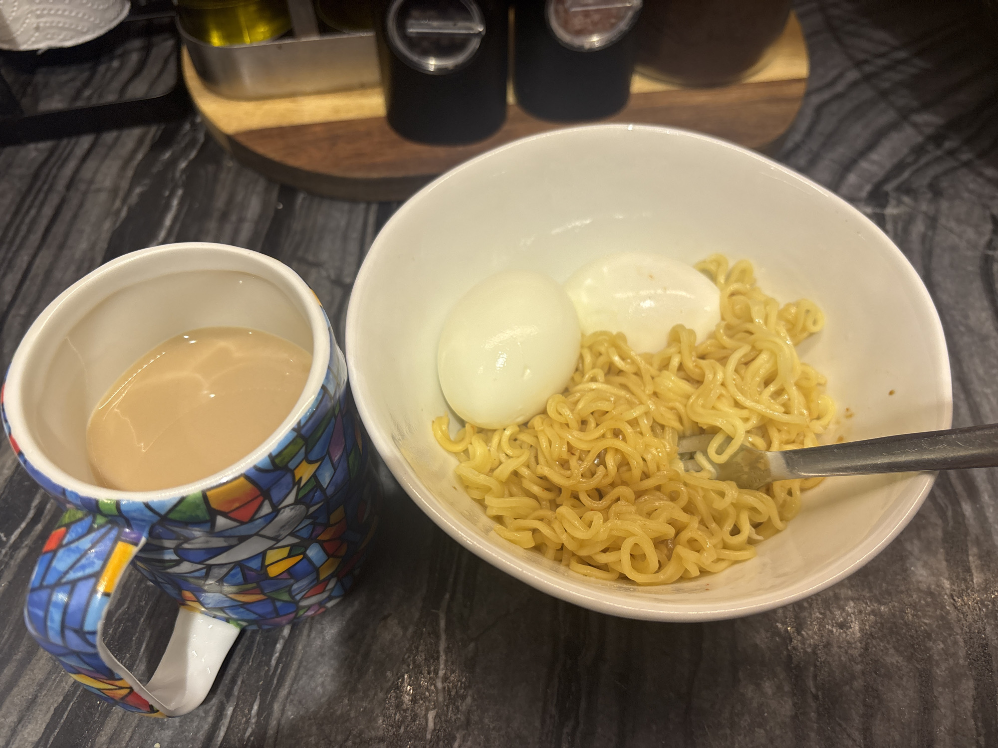 In this overhead shot of a dark countertop, a white bowl holds instant ramen noodles topped with two hard-boiled eggs. A fork is partially submerged in the noodles. To the left, a mug with a colorful stained-glass pattern contains a light brown beverage, possibly tea or coffee. The background shows a wooden cutting board and dark salt and pepper grinders, suggesting a kitchen setting. The scene does not provide any information about its location in Dubai, United Arab Emirates.