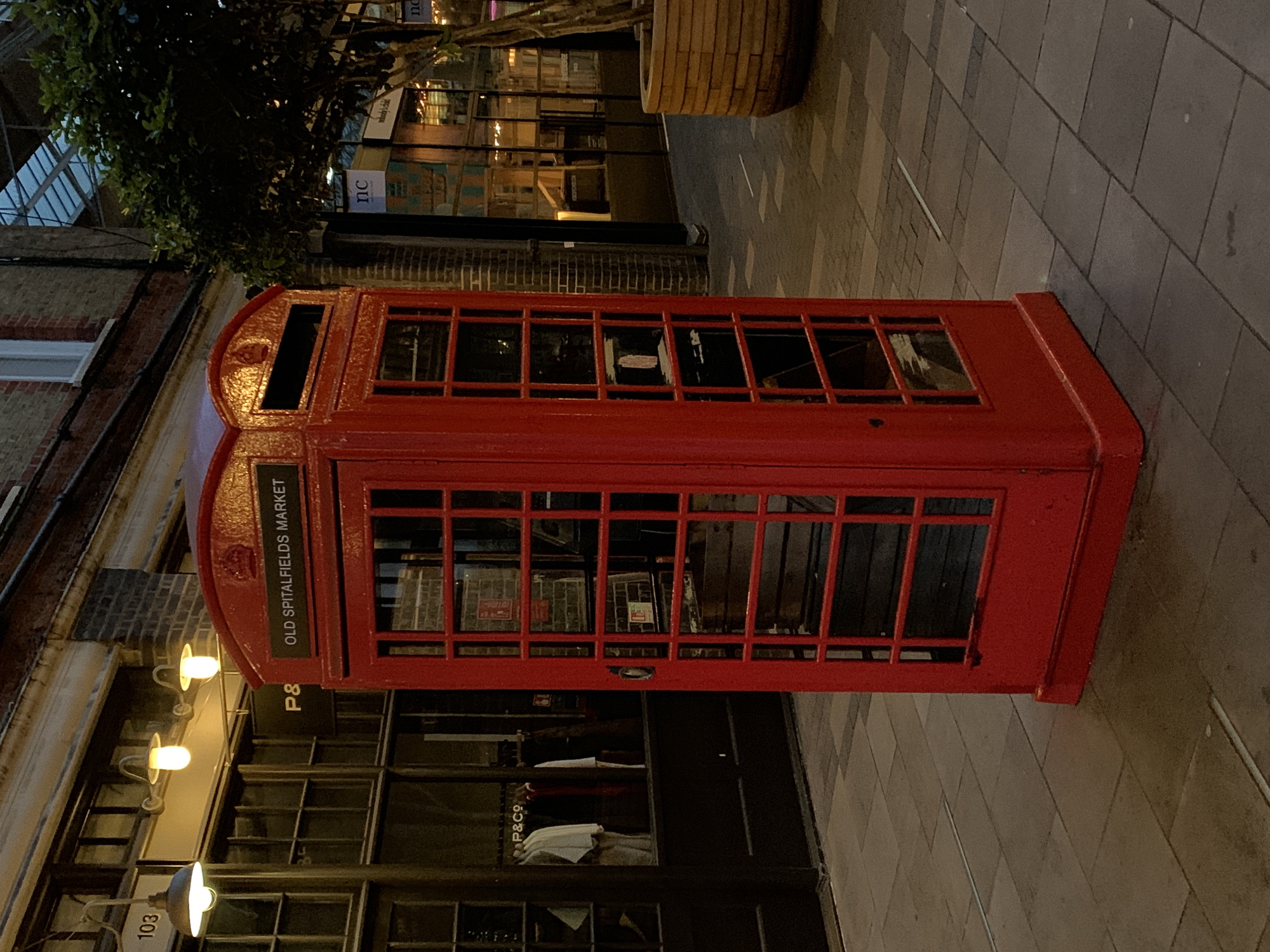 This image captures a classic red British telephone box (K6 kiosk design) standing on a paved pedestrian street in London, United Kingdom, specifically at Old Spitalfields Market. The photograph is taken in the evening or at night, indicated by the dark ambient light and the illumination from artificial sources such as street lamps and shop lights.
The iconic red telephone box is positioned prominently, showing signs of use but maintaining its vibrant colour. On its upper front panel, white text on a black background clearly spells out "OLD SPITALFIELDS MARKET," pinpointing the location. The telephone box has multiple glass panes, some of which reflect the surrounding lights.
To the left of the telephone box, buildings with brick facades and large glass shopfronts line the street. Warm, yellow light emanates from several street lamps mounted on the building walls, casting a glow on the immediate area. Above the buildings, dark green foliage from trees or large potted plants is visible against the night sky. On a shop window or sign, the text "P&CO" is visible twice, and other signs in the background display "TIC" and "nc." The building near the lamps also shows the number "103." The ground is paved with a pattern of rectangular and square tiles in grey and beige tones.
No people are present in the scene, which conveys a quiet and atmospheric sense of an urban market area after hours. The overall impression is one of a characteristic London street scene at night, highlighted by the historic telephone box.