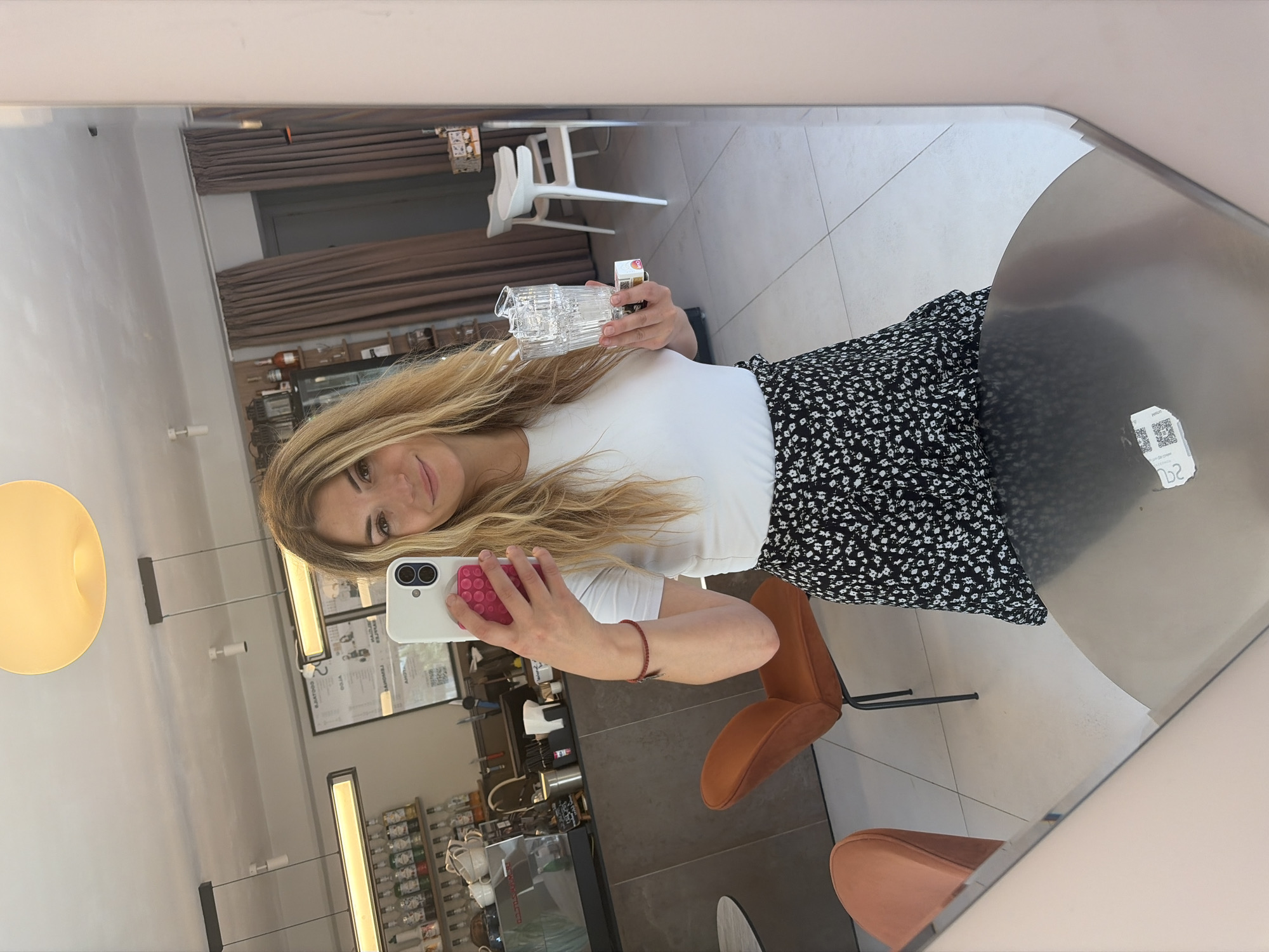 In Budva, Montenegro, a woman stands and takes a mirror selfie in what appears to be a cafe or bar. She is wearing a white t-shirt and a black and white floral skirt. She holds her smartphone in her right hand, capturing the reflection. In her left hand, she holds a clear glass and a small white object. The background shows a cafe interior with shelves of bottles, a coffee machine, seating, and a large mirror.