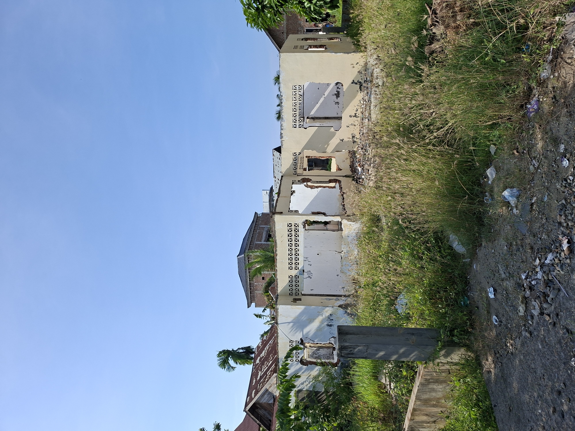 This outdoor scene, captured in Lhokseumawe, Indonesia, depicts a desolate landscape featuring several dilapidated buildings and overgrown vegetation under a clear, bright blue sky. The image is taken during broad daylight, indicated by the strong illumination and absence of clouds.
In the foreground and extending into the mid-ground, a rough dirt path or road runs along the right side, heavily littered with various debris including plastic bags, wrappers, and other trash. Adjacent to this path, and throughout much of the lower and central parts of the image, is a dense growth of tall, dry-looking grass and weeds, interspersed with some greener foliage, also showing signs of litter.
The main subjects are a series of abandoned or severely damaged single and possibly two-story buildings, appearing to be constructed of concrete or stucco. Their walls, primarily a faded light yellow or cream color, are crumbling in many places, revealing exposed brickwork or bare concrete. Windows are empty frames, and several openings show the interiors of the structures. Some upper wall sections feature decorative concrete ventilation blocks with geometric, 'X'-like patterns. The roofs are either partially missing or in poor condition, with one structure showing a rusted corrugated metal roof. Lush green trees and palm fronds are visible around and behind these buildings.
No people are visible, and no specific event is taking place, contributing to a sense of abandonment and neglect. A concrete pillar stands upright in the middle ground, near the buildings and vegetation. There is no legible text visible within the frame. The overall impression is one of urban decay or post-disaster neglect in a tropical environment.