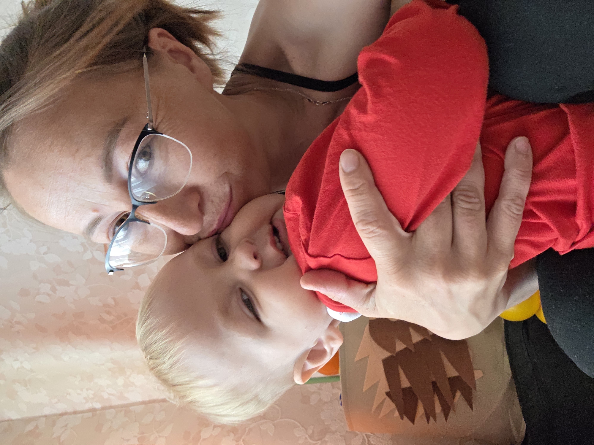A woman wearing glasses and a black choker necklace is holding a young child dressed in a red garment. The child is angled towards the camera, with a hand holding onto the red fabric. The background includes a surface with a floral pattern and a cardboard container. The location is Unknown, Unknown.