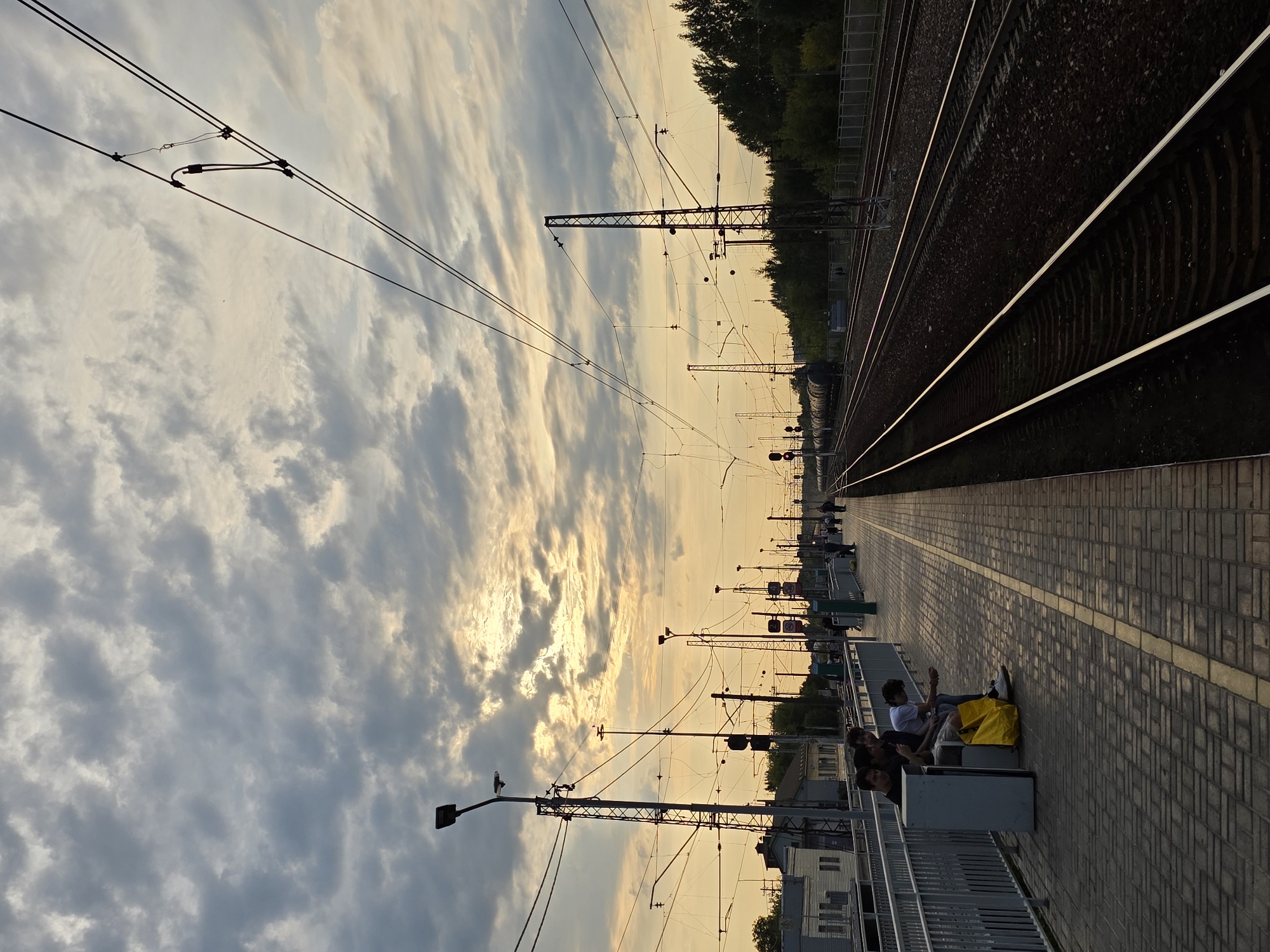 The image depicts a train station platform at dusk in Posolok Kombinata Stroymaterialov-2, Russia. The sky is filled with dramatic, textured clouds illuminated by the setting sun. Several individuals are present on the platform, with a group of four young men sitting on a bench. One of them is holding a yellow bag. Overhead, a complex network of electrical wires for the trains crisscrosses the sky, supported by tall metal poles. The platform itself is paved with brick-like tiles and has a railing along the edge. Adjacent to the platform are multiple sets of train tracks, stretching into the distance and bordered by trees and vegetation on the right. Buildings are visible in the background on the left side of the image.