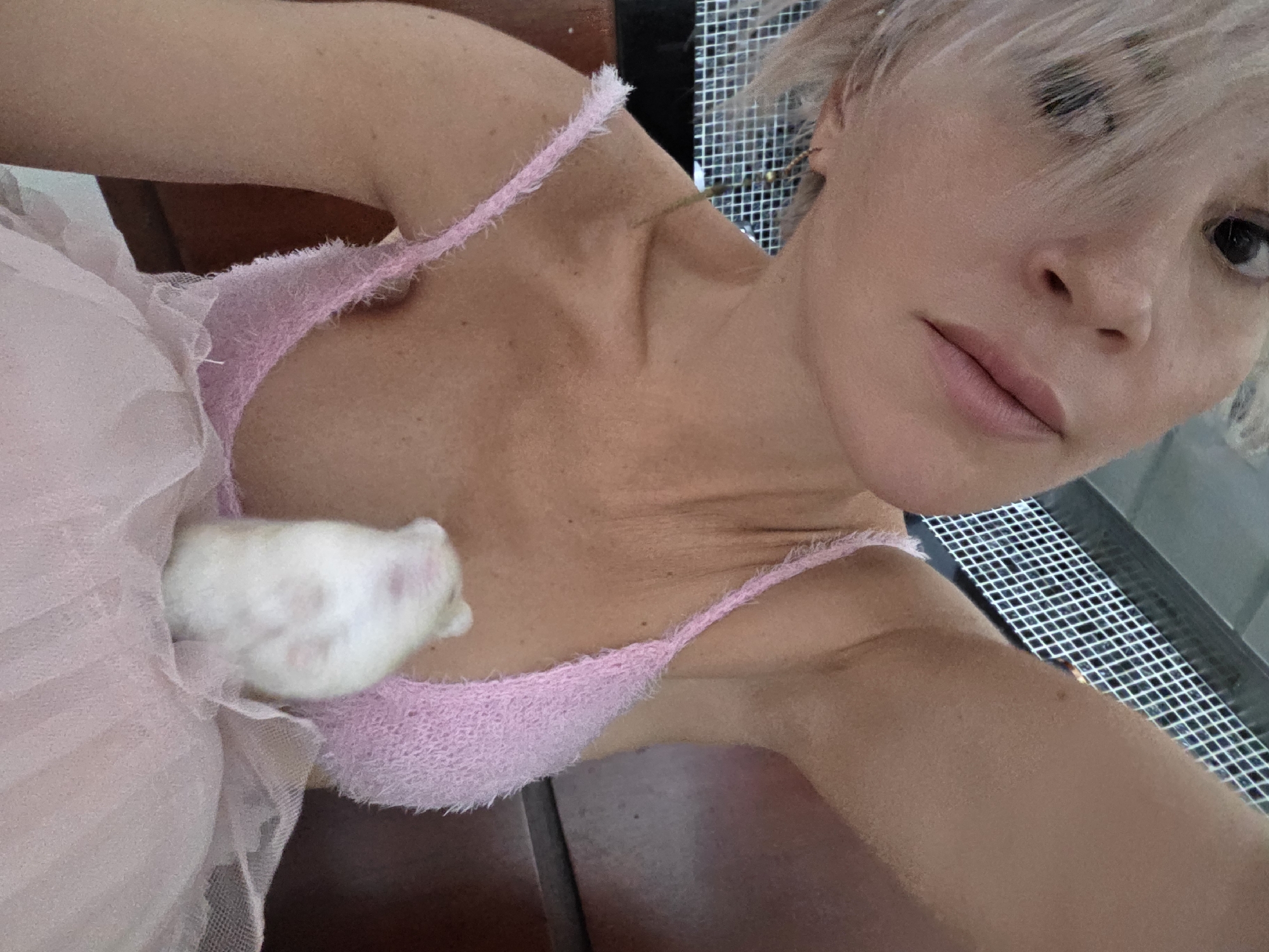A close-up, high-angle shot shows a woman with short, blonde hair, her face angled away from the camera. She is wearing a pink, fuzzy bralette and a light pink tulle skirt. A small, white and pink hamster is nestled in the cleavage of her bralette, peeking out from the top of the fabric. The background appears to be indoors, with a metal grate and wooden surfaces visible. The scene is set in a private residence in North Kuta, Indonesia.