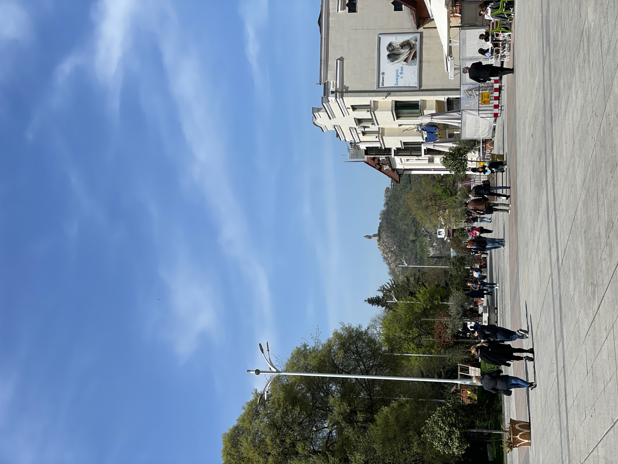 This image captures a vibrant pedestrian street scene in Plovdiv, Bulgaria, under a clear blue sky with sparse, wispy clouds. The perspective looks down a wide, paved walkway lined on one side by buildings and on the other by lush greenery.
On the left side of the image, numerous green trees, some showing fresh spring foliage, border the street. A modern street lamp with three arched fixtures stands prominently in the foreground. In the mid-ground, a rocky, tree-covered hill rises, with a small structure visible at its peak, suggesting a distant landmark or historical site.
The right side of the street features multi-story buildings with various architectural details and windows. Several outdoor cafes or restaurants are present, indicated by awnings and tables with people seated. A prominent billboard on one building displays the "Allianz" logo and the Cyrillic text "Заедно с Вас," which translates to "Together with You," accompanied by an image of a person resting their head.
The paved street in the foreground and mid-ground is bustling with people. Individuals and small groups are seen walking, strolling, and sitting at the outdoor establishments. They are dressed in casual clothing, suitable for pleasant, sunny weather. A red and white striped barrier is visible near one of the cafes, possibly indicating a temporary obstruction or sectioned-off area.
The time of day appears to be bright daylight, possibly late morning or afternoon, given the clear visibility and evident shadows. The overall atmosphere is lively and relaxed, depicting a typical day in a popular urban area.