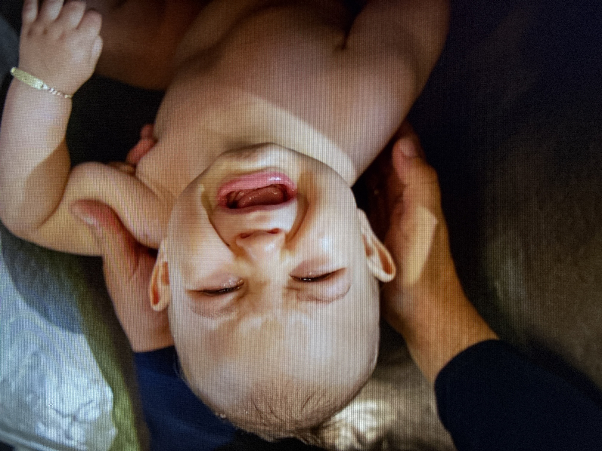 An overhead close-up shot captures a baby being held in what appears to be a bathing environment. The baby, an infant or young toddler with fair skin and some dark hair visible on the back of its head, is crying or screaming. Its mouth is wide open, revealing its tongue, and its eyes are squeezed shut, conveying clear distress. Water droplets are visible on the baby's face, suggesting it is in water, possibly a bathtub or basin.
Two adult hands are visible, supporting the baby securely. One hand holds the baby's left arm and shoulder, while the other supports the baby's head near its right ear. A small, light-colored bracelet is worn on the baby's left wrist. The adult's right hand has a dark blue sleeve, possibly from a shirt or jacket. The background is dark and somewhat reflective, consistent with a wet, enclosed space. The scene depicts the baby experiencing acute discomfort during an activity like bathing. No specific time of day or weather cues are present. The photo was taken in Varna, Bulgaria.