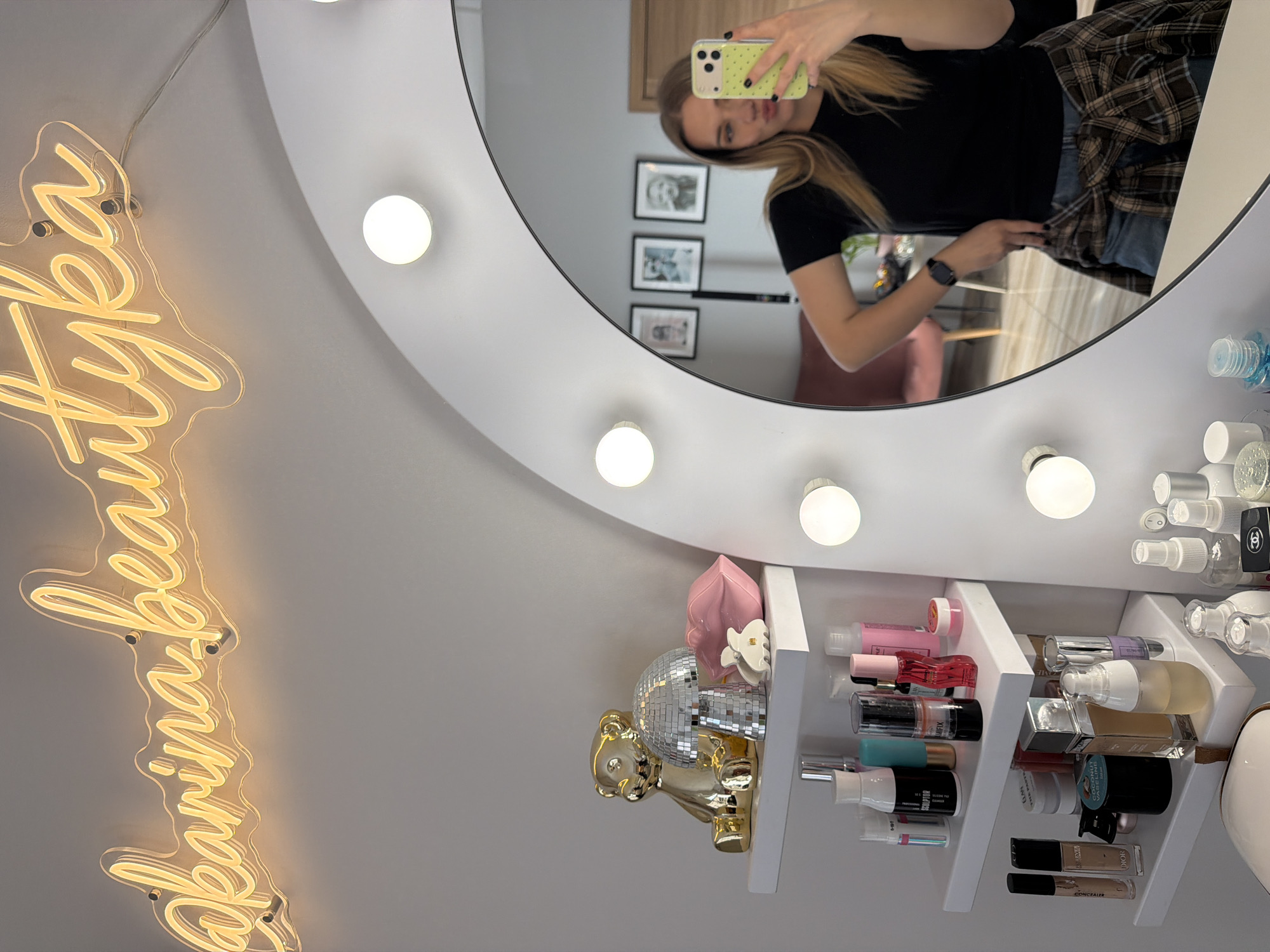 The image shows a vanity mirror with lights and shelves filled with beauty products. A person is visible in the reflection, holding a phone, presumably taking a selfie. On the shelves, there are various cosmetic items like perfumes, lotions, and makeup. There's a decorative golden bear and a disco ball on the left side of the shelves. On the wall, a neon sign reading "@karinabeautyka" is mounted.
While it's impossible to determine the exact location in Riga, Latvia from the image, the social media handle in the neon sign suggests the vanity belongs to a beauty influencer or business operating under the name "karinabeautyka". The specific address or region within Riga remains unknown.